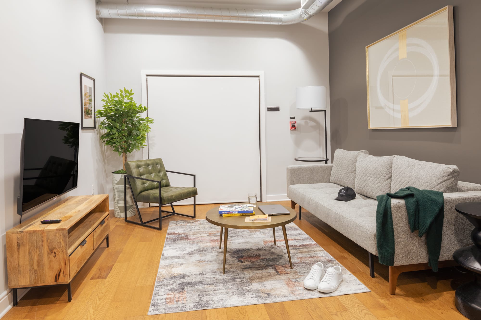 A cozy living room with a light grey sofa, a green leather armchair, and a wooden TV stand with a flat-screen TV. A large abstract artwork hangs above the sofa, and a tall potted plant sits in the corner.