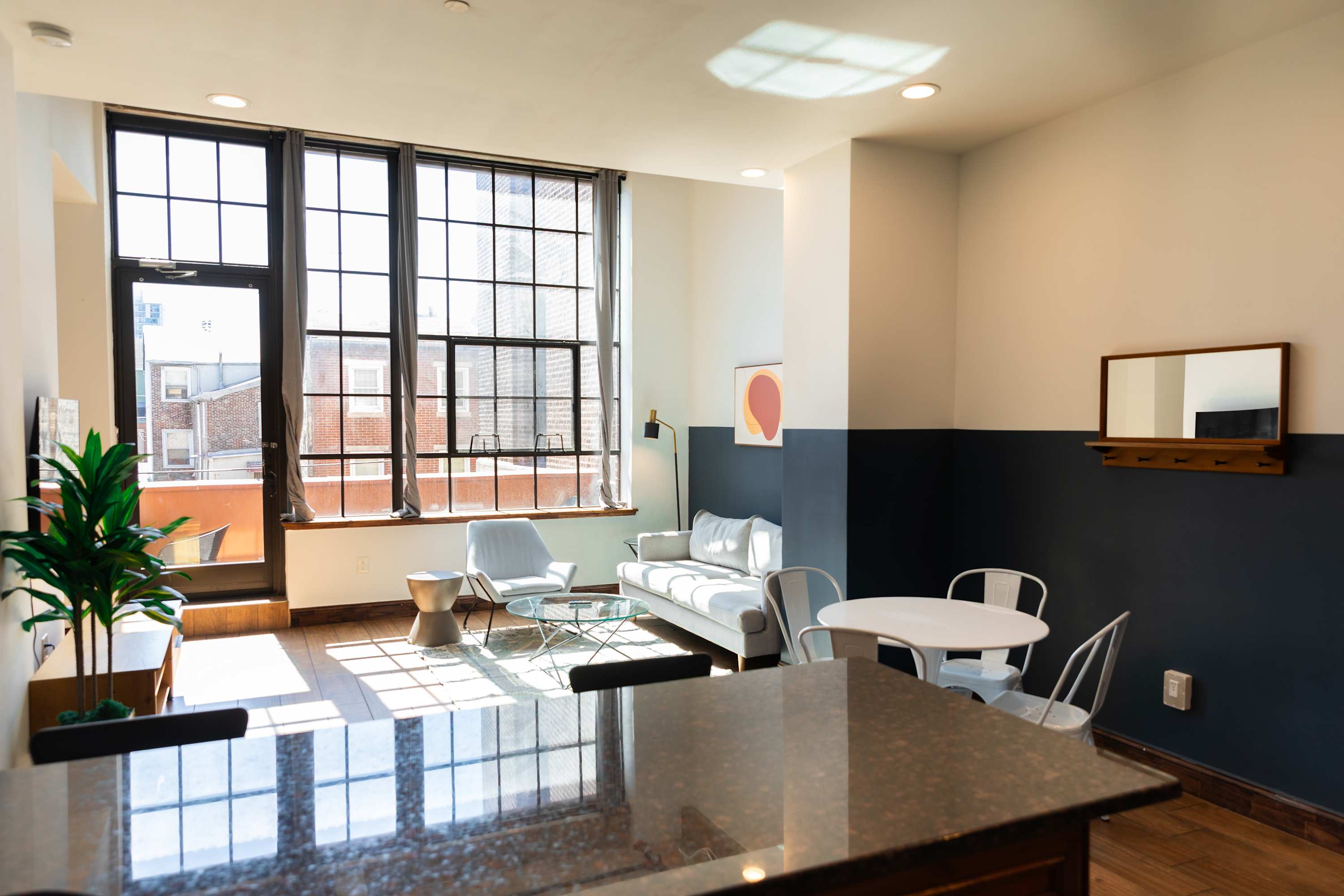 Spacious living area with large industrial-style windows and a glass door leading to a balcony, featuring a light grey sofa, two armchairs, a glass coffee table, and a dining area with a round white table and chairs, all on hardwood floors.