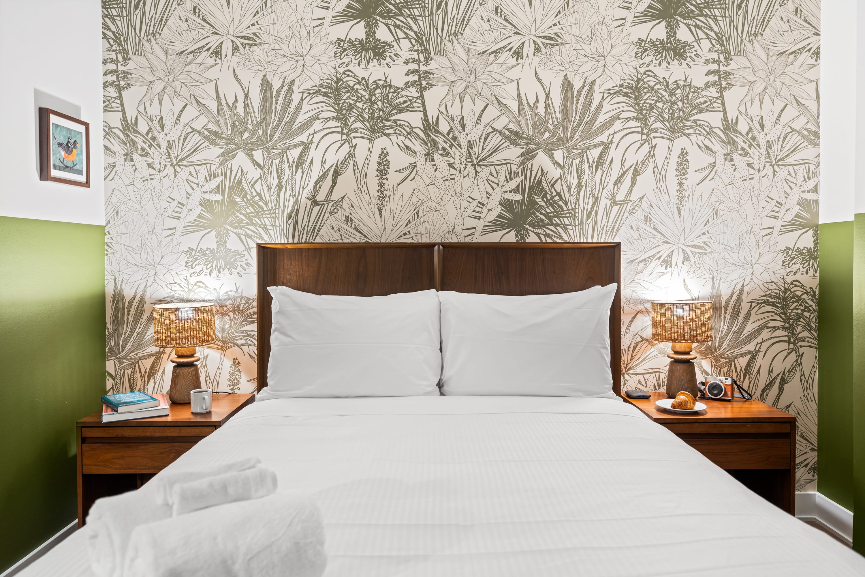 Cozy bedroom featuring a dark wood bed frame with white bedding, flanked by two nightstands with woven lamps, and a striking tropical-patterned wallpaper accent wall.