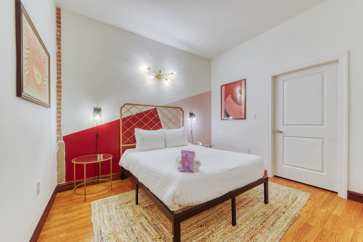 A stylish bedroom with a queen-sized bed featuring a gold geometric headboard, set against a bold accent wall painted with red and pink geometric shapes, and illuminated by a modern sputnik chandelier.