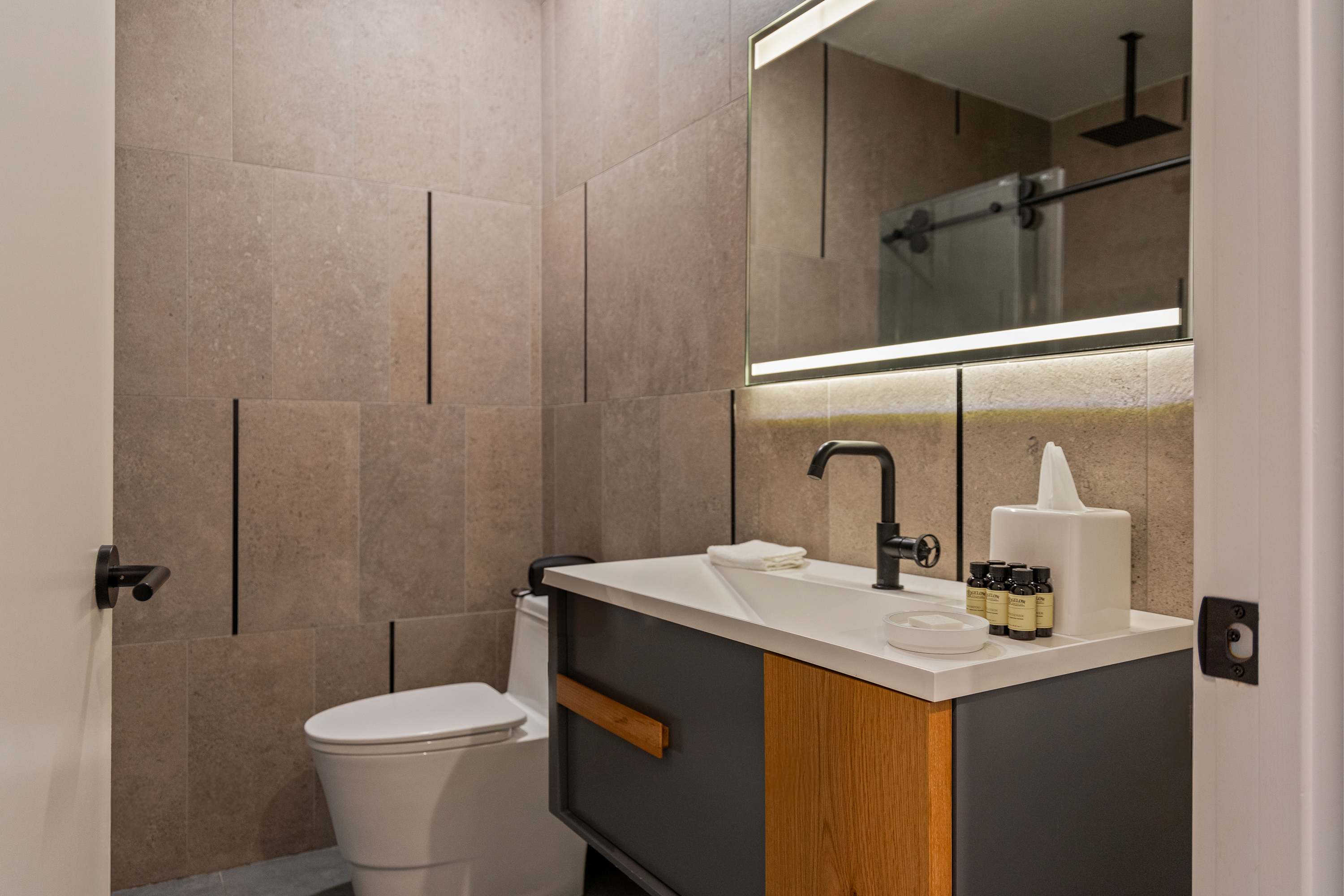 Contemporary bathroom with light brown vertical tile walls and a modern vanity featuring a dark gray cabinet with a wood accent, a white countertop, and a sleek black faucet. A large mirror with integrated lighting is positioned above the vanity.