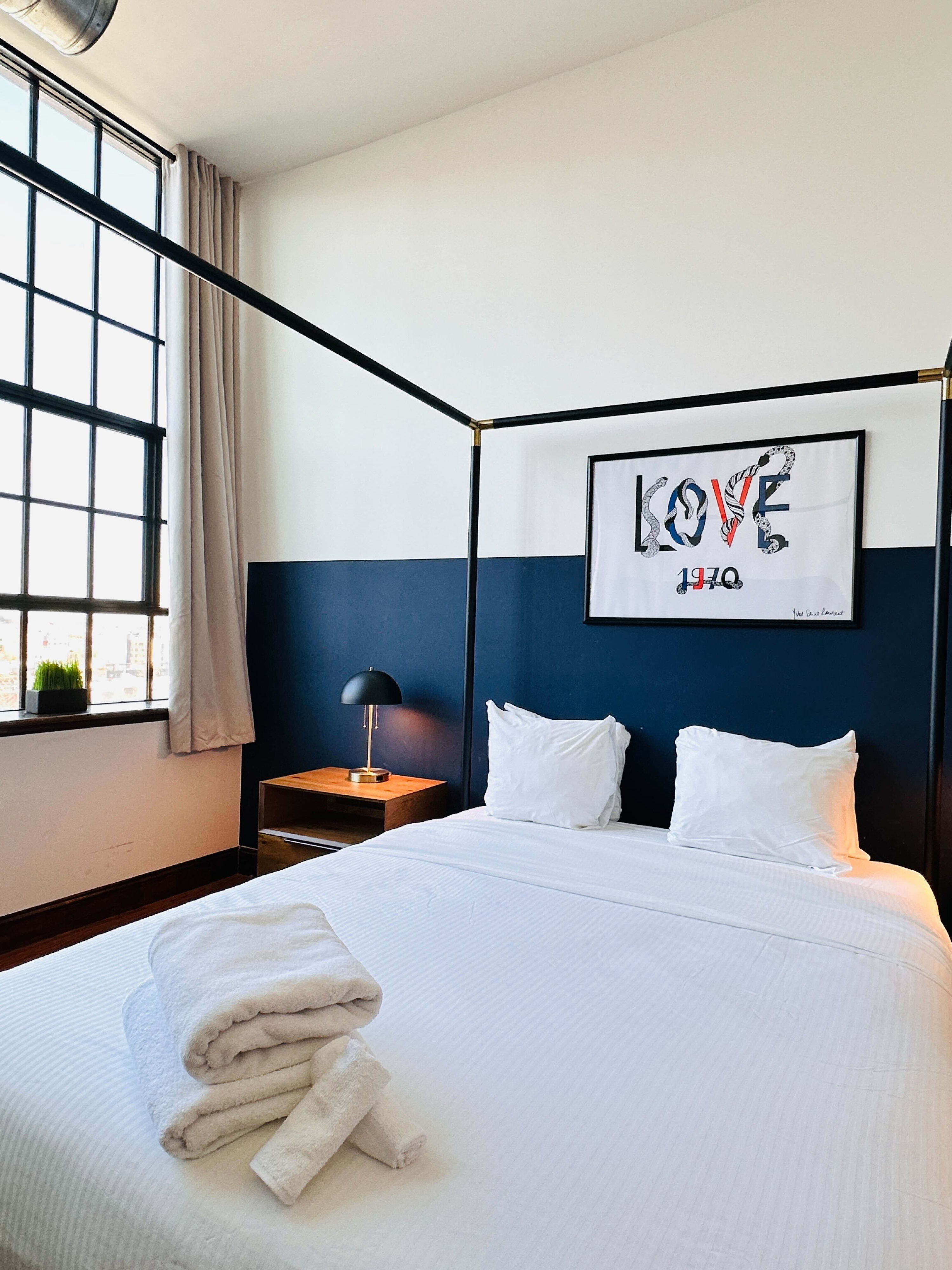 Close-up view of the black four-poster bed with crisp white bedding and neatly folded white towels. A wooden nightstand with a black lamp is visible, and a large window with city views is in the background.