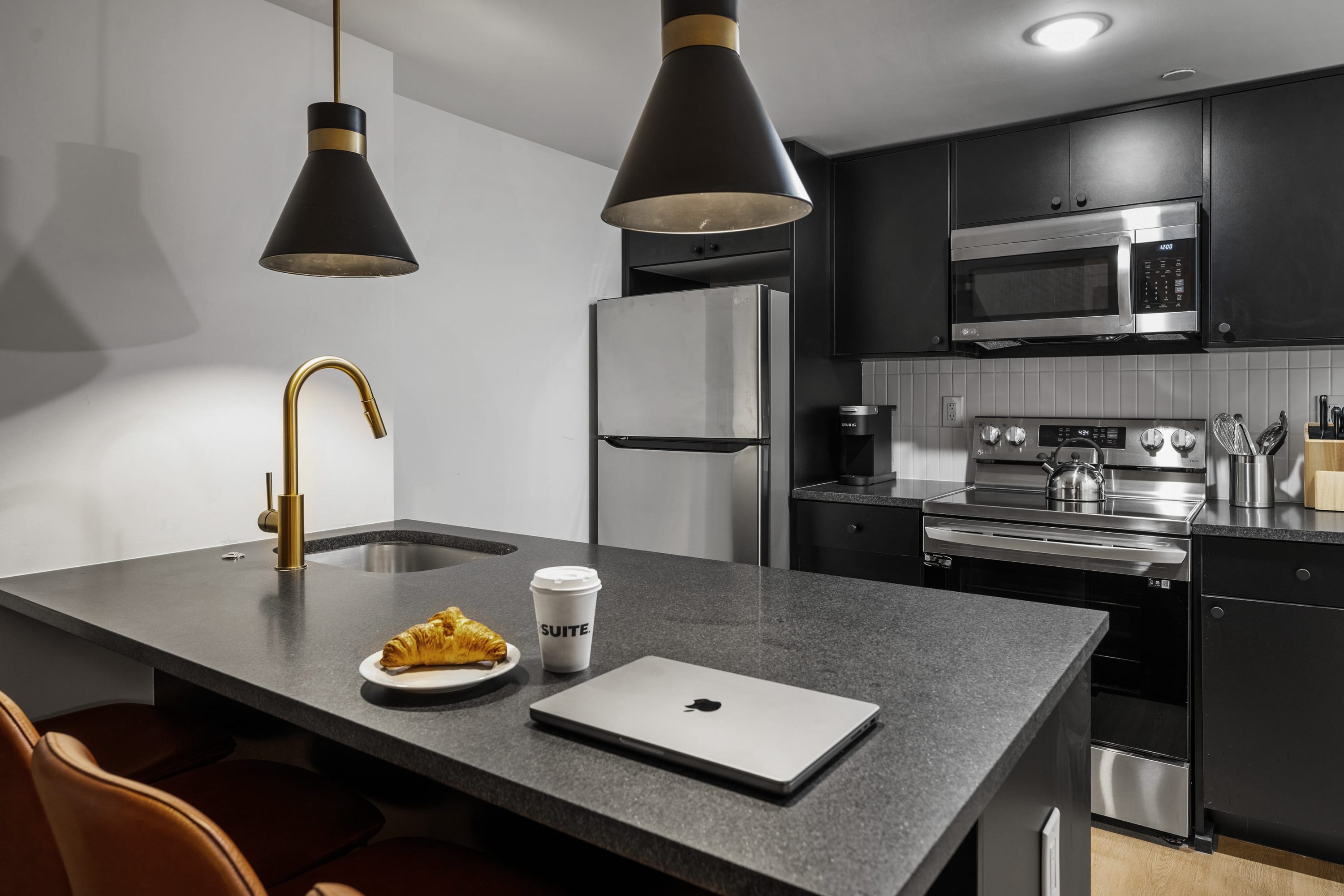 Modern kitchen with dark gray cabinetry, stainless steel appliances, and a large island with a dark countertop and gold faucet. Two black pendant lights hang above the island, which holds a laptop, croissant, and coffee cup.