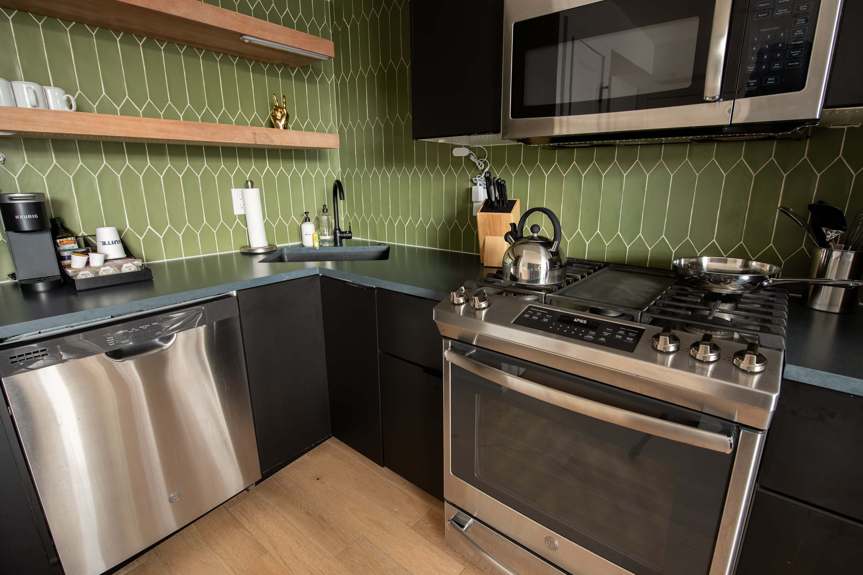 A modern kitchen featuring dark lower cabinets, a vibrant green picket tile backsplash, stainless steel appliances including a dishwasher, oven range, and microwave, complemented by open wooden shelving.