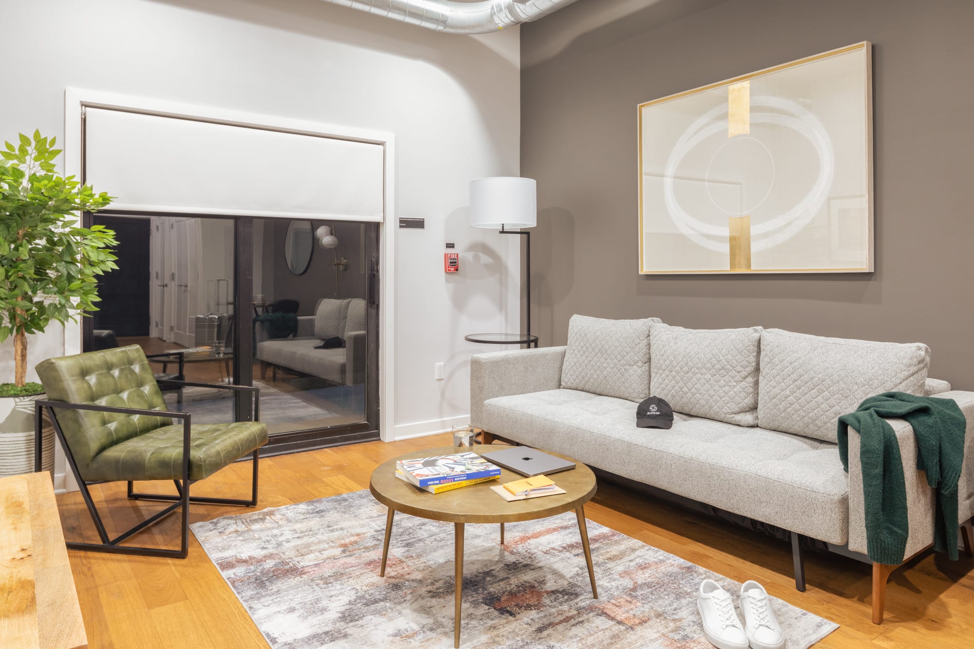 A contemporary living room with a light grey sofa, a green leather armchair, and a round brass coffee table on a patterned rug. A large glass door leads to an outdoor space, and a tall plant adds a touch of greenery.