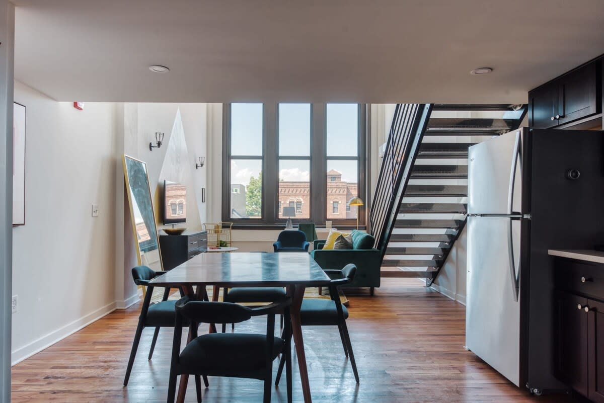 Dining area with a dark rectangular table and four chairs, adjacent to a modern kitchen with dark cabinets and stainless steel appliances. The view opens to the living room with large windows.