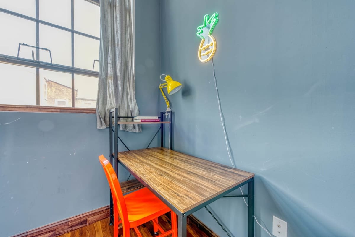 A dedicated workspace within a bedroom, featuring a wooden desk with a black frame, a bright orange chair, a yellow desk lamp, and a playful pineapple neon sign on the blue-grey wall next to a large window.