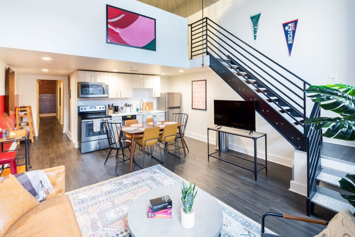 Bright open-concept loft featuring a modern white kitchen with stainless steel appliances, a wooden dining table, and a living area with a tan sofa and patterned rug. A black metal staircase ascends to an upper floor.