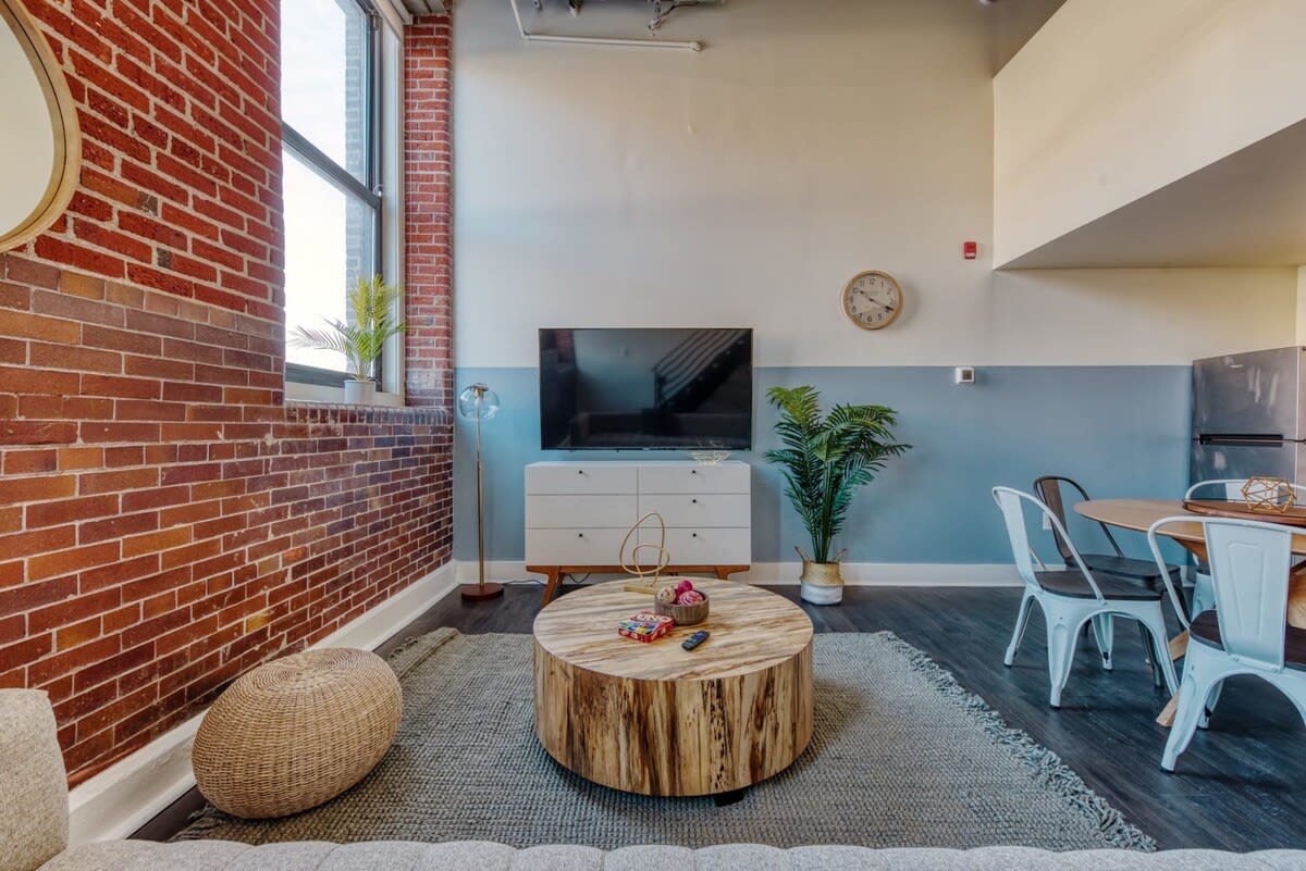 Stylish living room with an exposed brick wall and large windows, featuring a light gray tufted sofa, a round wooden coffee table on a textured rug, and a flat-screen TV on a white console. A potted plant adds a touch of greenery.