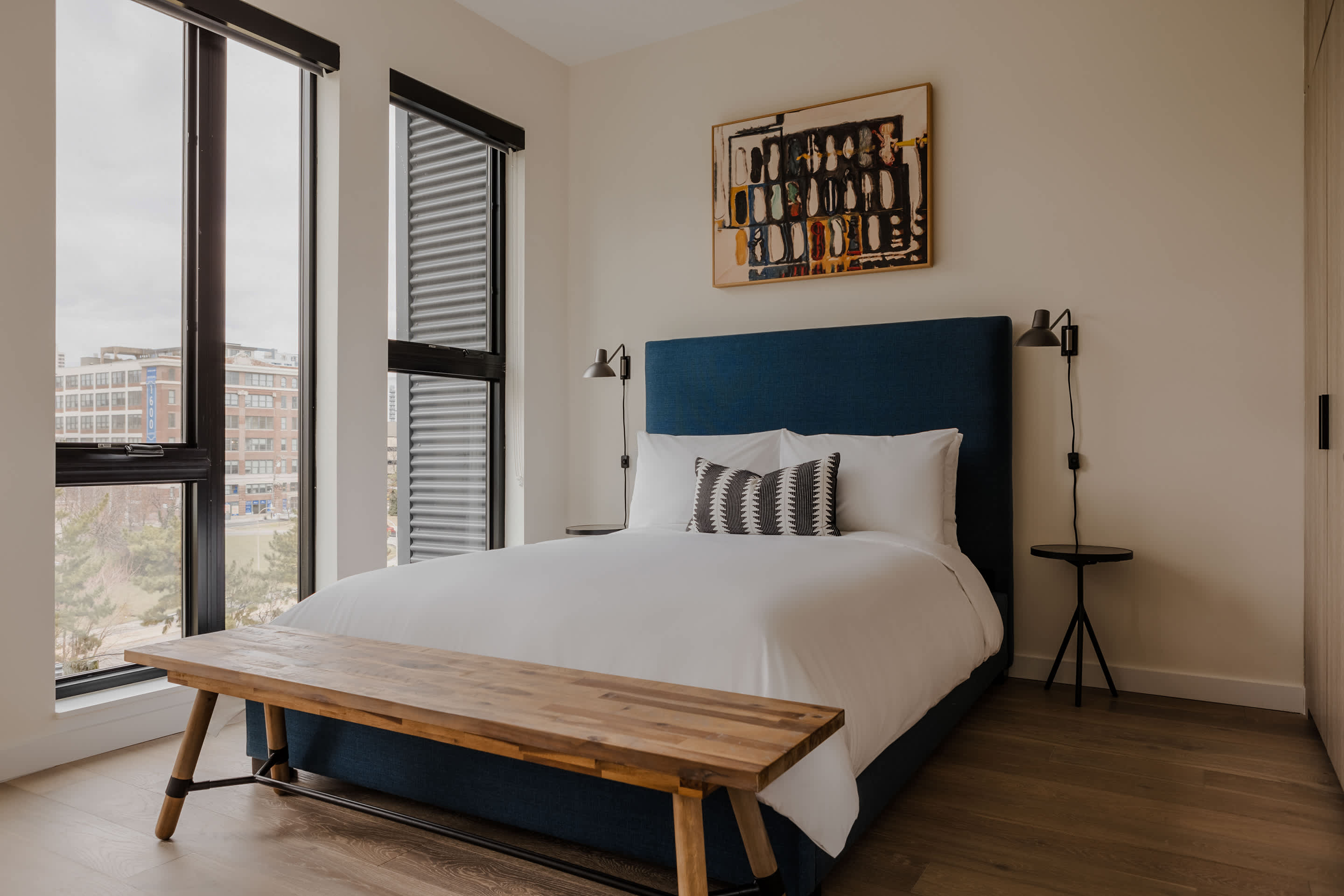 Contemporary bedroom with a blue upholstered headboard, crisp white bedding, and a natural wood bench at the foot of the bed, flanked by black wall-mounted lamps and large windows.