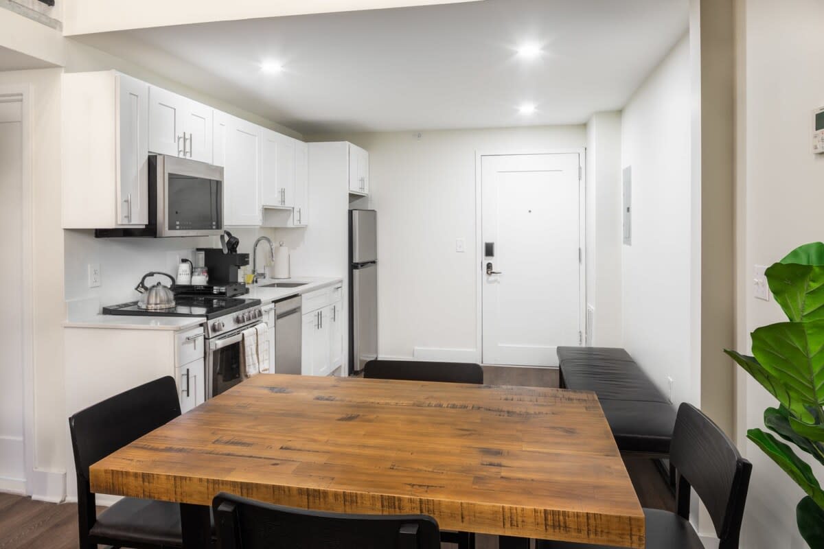 Cozy dining area with a rustic wooden table and black chairs, positioned next to a modern kitchen with white cabinets and stainless steel appliances, with the main entrance door visible.