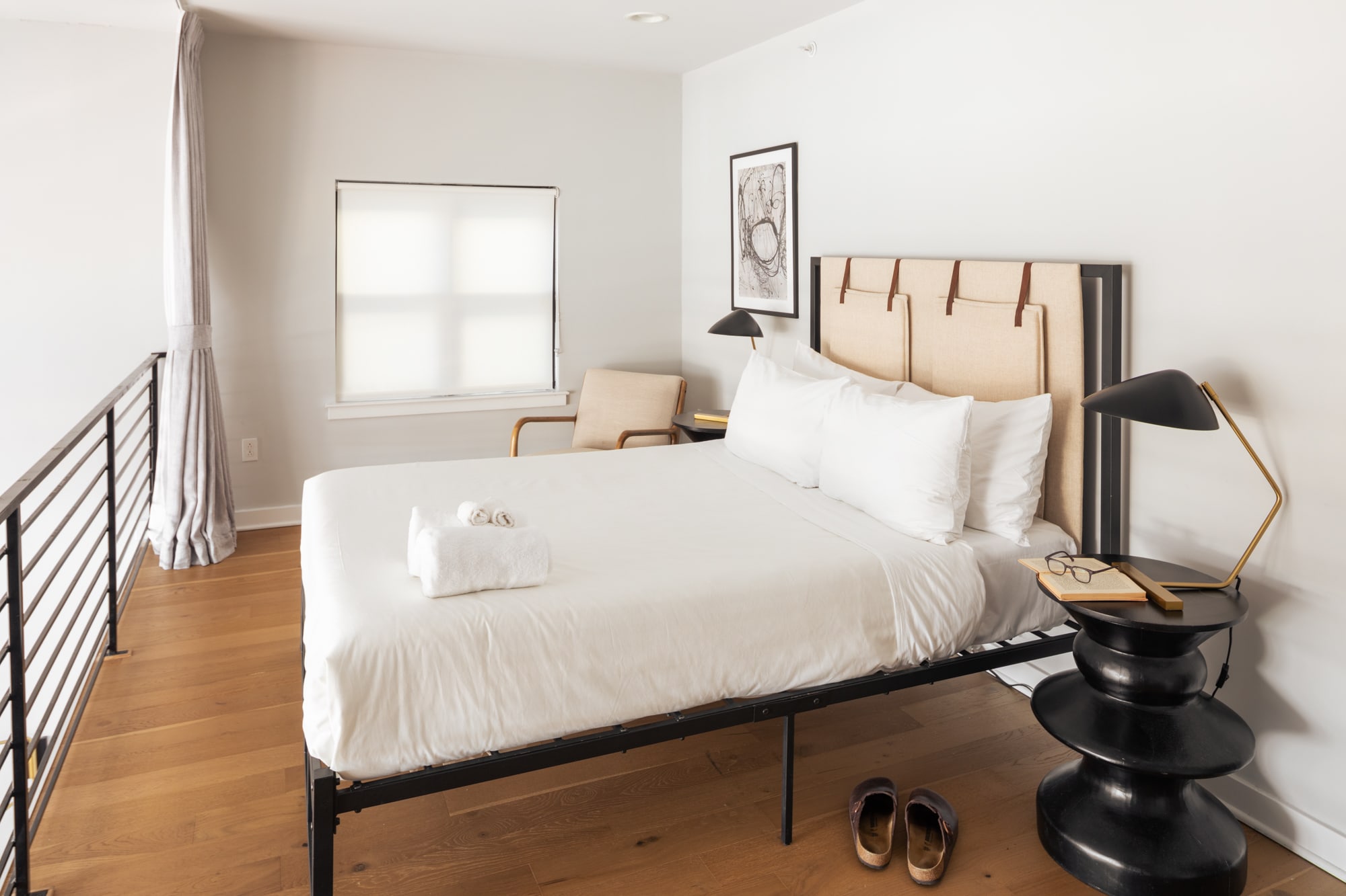 A bright bedroom with a queen-sized bed featuring a black metal frame and a beige upholstered headboard, light hardwood floors, a black tiered nightstand with a lamp, and a small armchair by a window with white roller shades.