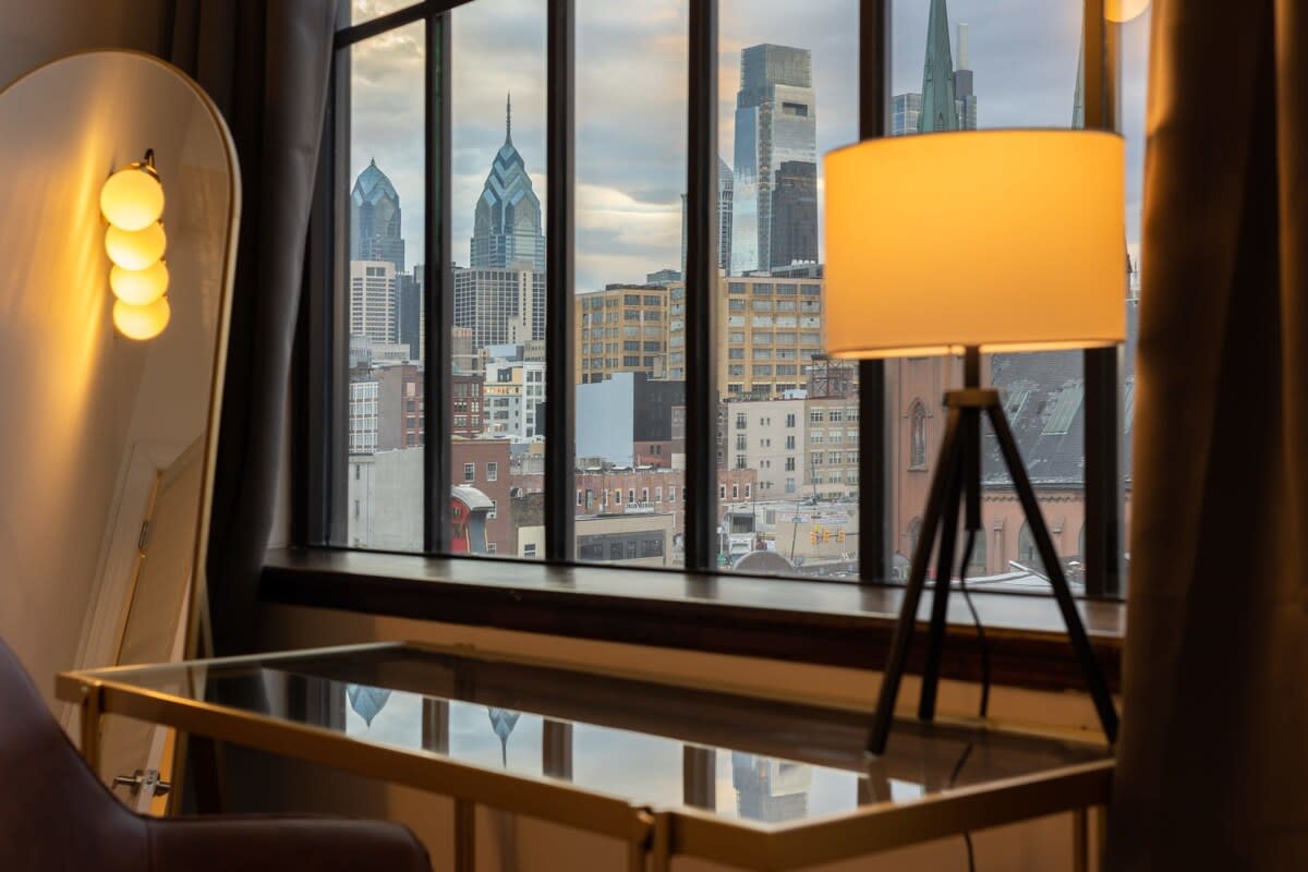 A close-up view of a city skyline with prominent skyscrapers, seen through large industrial-style windows. A modern desk lamp with a white shade and a tall, arched mirror with spherical lights are visible in the foreground.