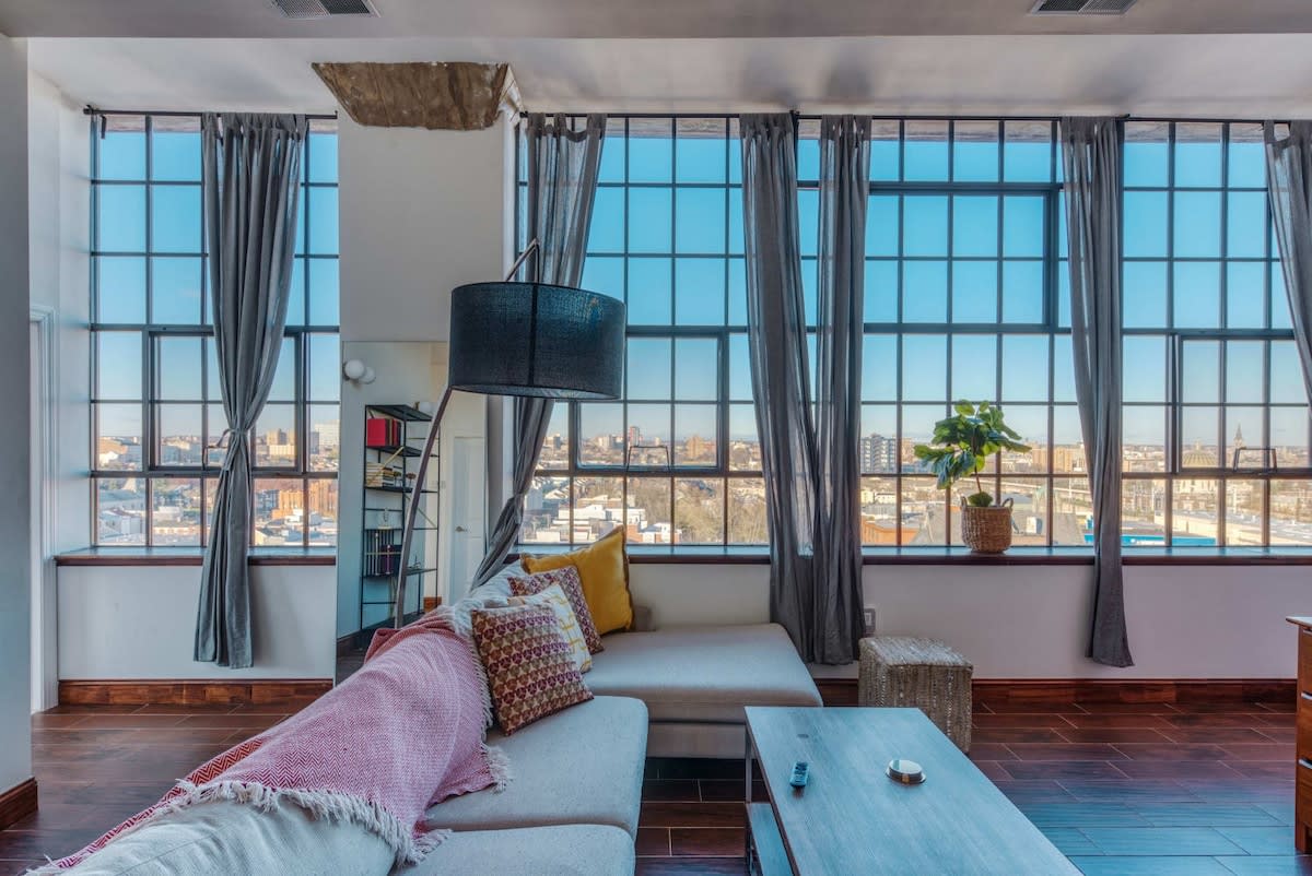 Bright living room with a large light-colored sectional sofa adorned with colorful pillows and a pink throw, facing expansive industrial-style windows with city views. Dark grey curtains frame the windows.