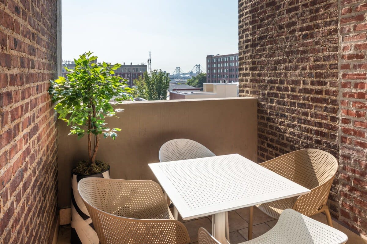 A cozy outdoor balcony enclosed by exposed brick walls, furnished with a white square table and four light-colored woven chairs. A potted green plant adds a natural touch, and a distant city skyline with a bridge is visible under a clear sky.