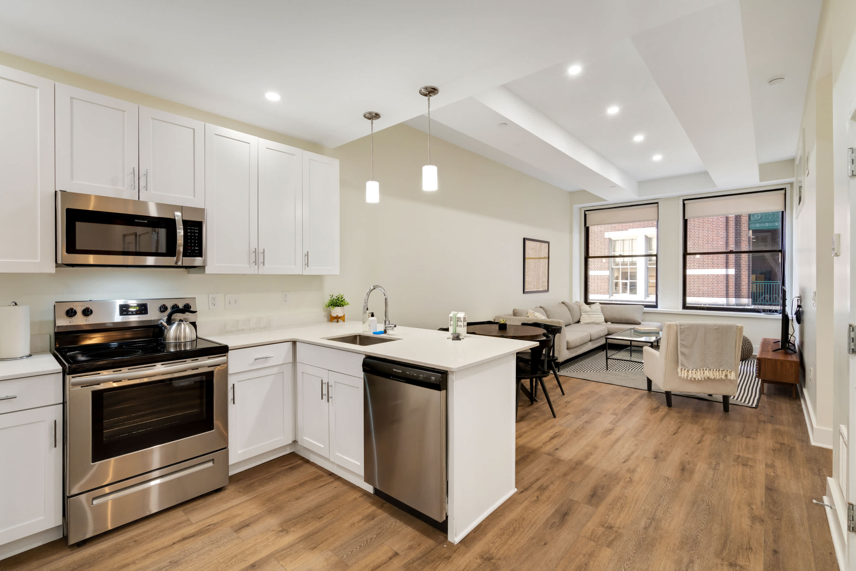 Open concept kitchen and living area with white shaker cabinets and stainless steel appliances, including a dishwasher. A round dining table with black chairs is visible in the foreground, leading to a living area with a sectional sofa and large windows.