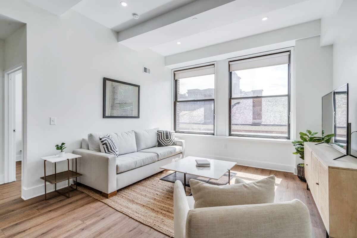Bright living room with a light gray sofa, a white marble coffee table, and a light-colored armchair, all resting on a textured jute rug. A large framed artwork hangs on the wall, and two tall windows provide natural light.
