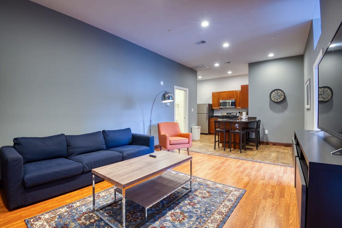 An open-concept living area with light hardwood floors transitioning to wood-look tile in the adjacent kitchen. The living room features a dark blue sofa, an orange armchair, and a wooden coffee table on a patterned rug, while the kitchen includes cherry wood cabinets and a breakfast bar with stools.