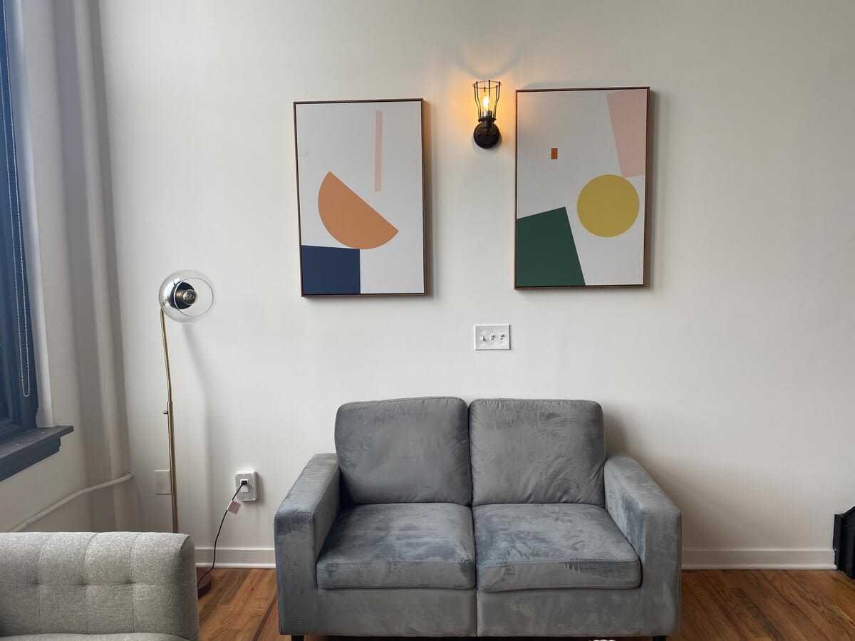 Section of the living room with a small gray loveseat, two abstract art pieces on the wall, and a modern floor lamp.
