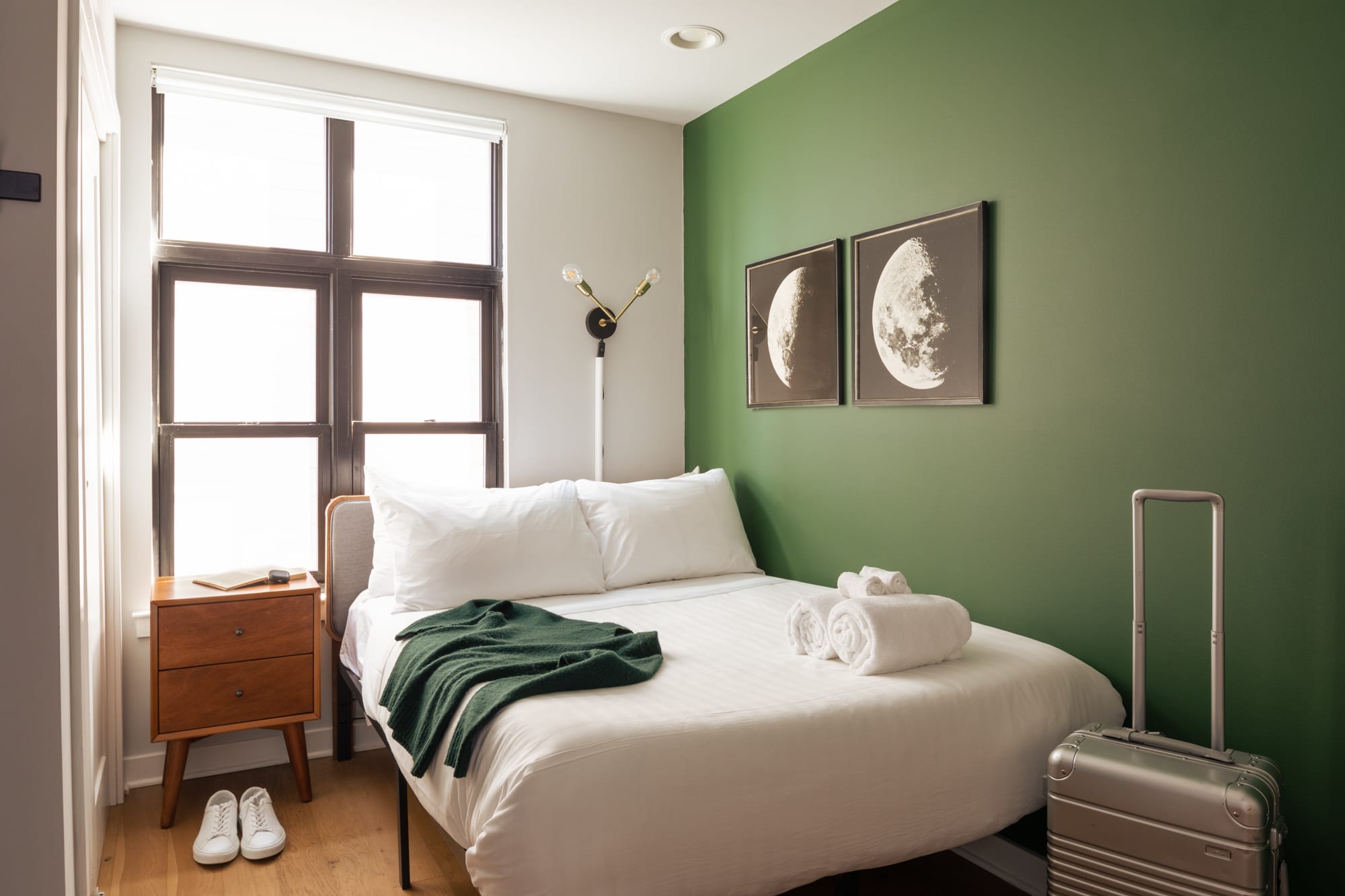 A cozy bedroom with a queen-sized bed, white bedding, a dark green accent wall, two moon-themed artworks, a wooden nightstand, and a silver suitcase at the foot of the bed.