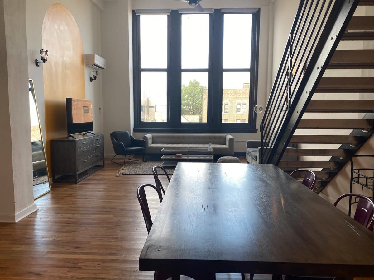 Wide view of the open-concept living and dining area with high ceilings, large black-framed windows, and a dark wooden dining table with purple metal chairs in the foreground. A staircase with metal railings leads upstairs.
