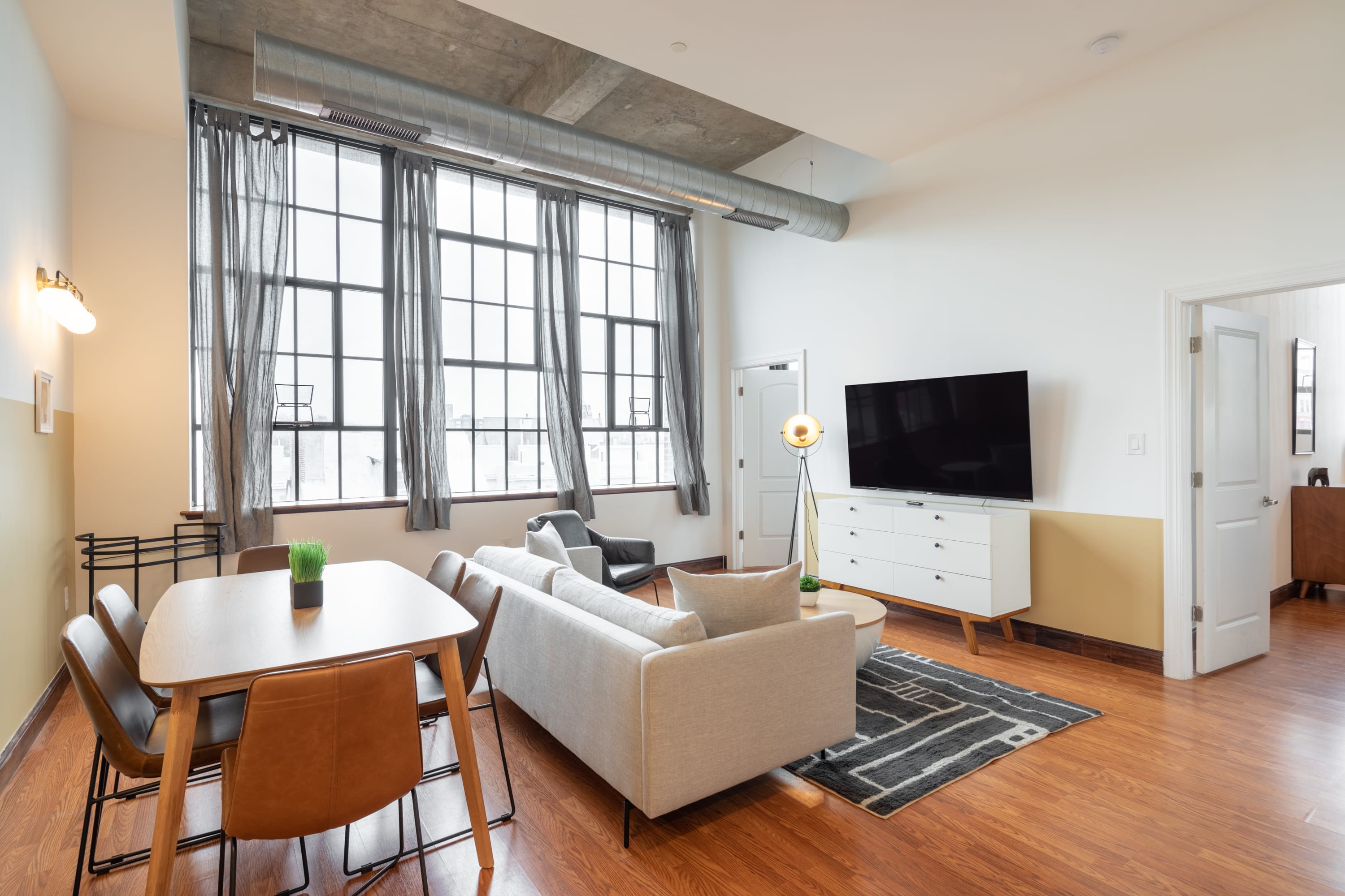 Spacious open concept living and dining area with a light gray sofa, a wooden dining table with brown chairs, and large industrial windows with gray curtains. A flat-screen TV is mounted above a white dresser, and a floor lamp adds a modern touch.