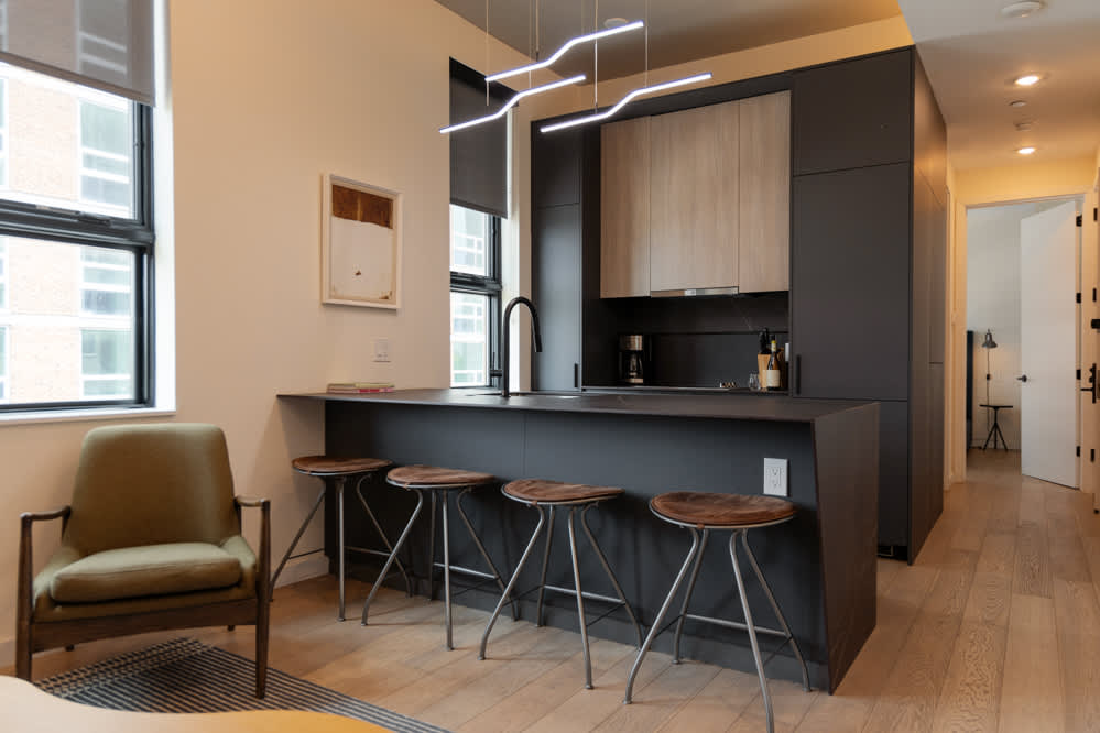 A modern open-concept kitchen features dark cabinetry and a large dark island with four wooden bar stools, illuminated by a contemporary linear chandelier. An olive green armchair sits on a striped rug in the foreground, connecting to the living space.