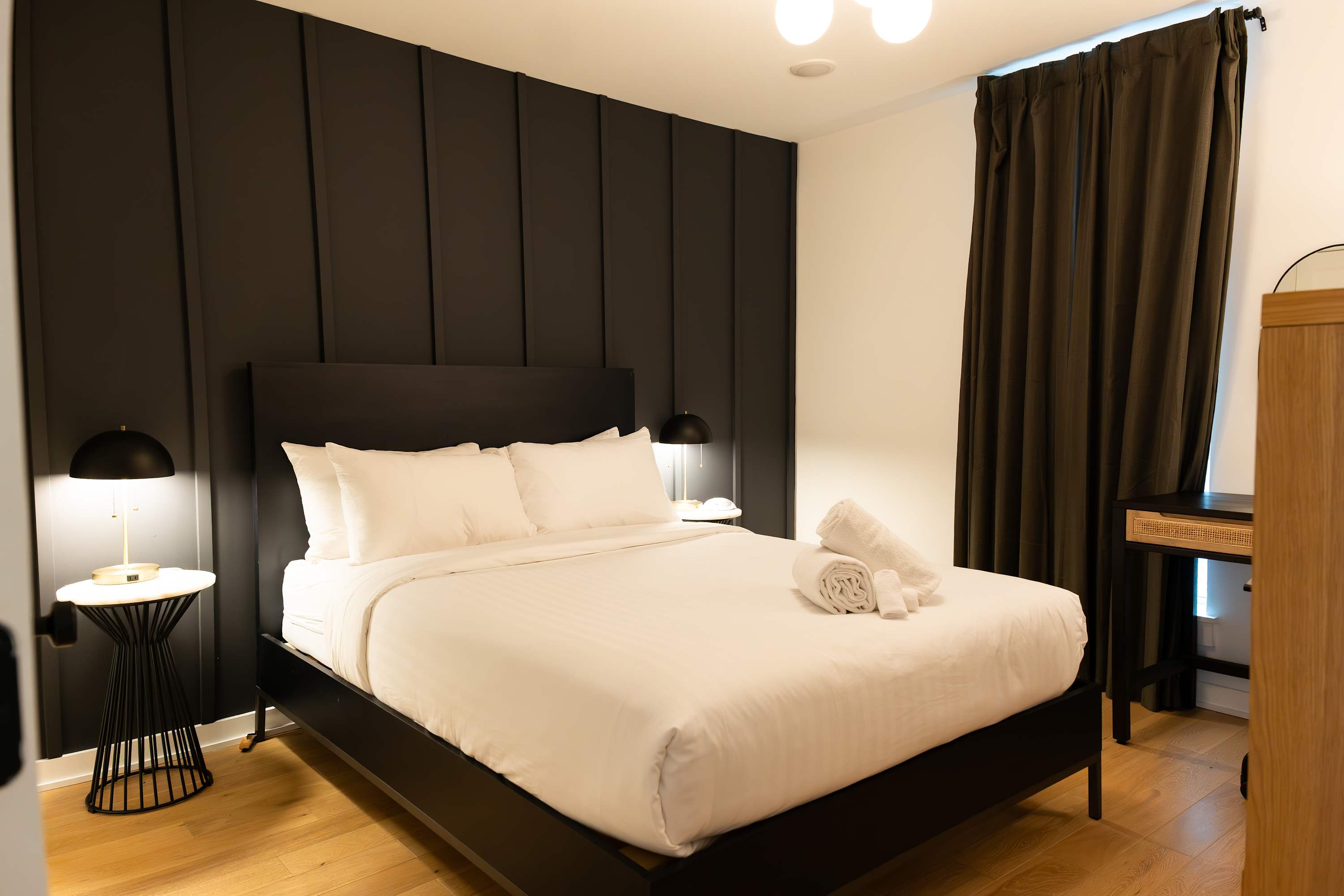 A stylish bedroom with a black paneled accent wall behind a queen-sized bed dressed in crisp white linens, flanked by modern black and gold nightstands with matching lamps. Dark curtains cover a window on the right.