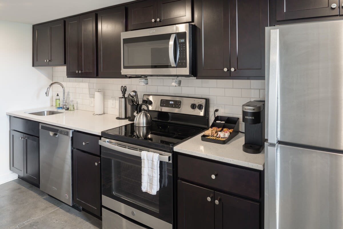 A modern kitchen with dark wood cabinets, white subway tile backsplash, and light quartz countertops. Stainless steel appliances include a microwave, range, dishwasher, and refrigerator.
