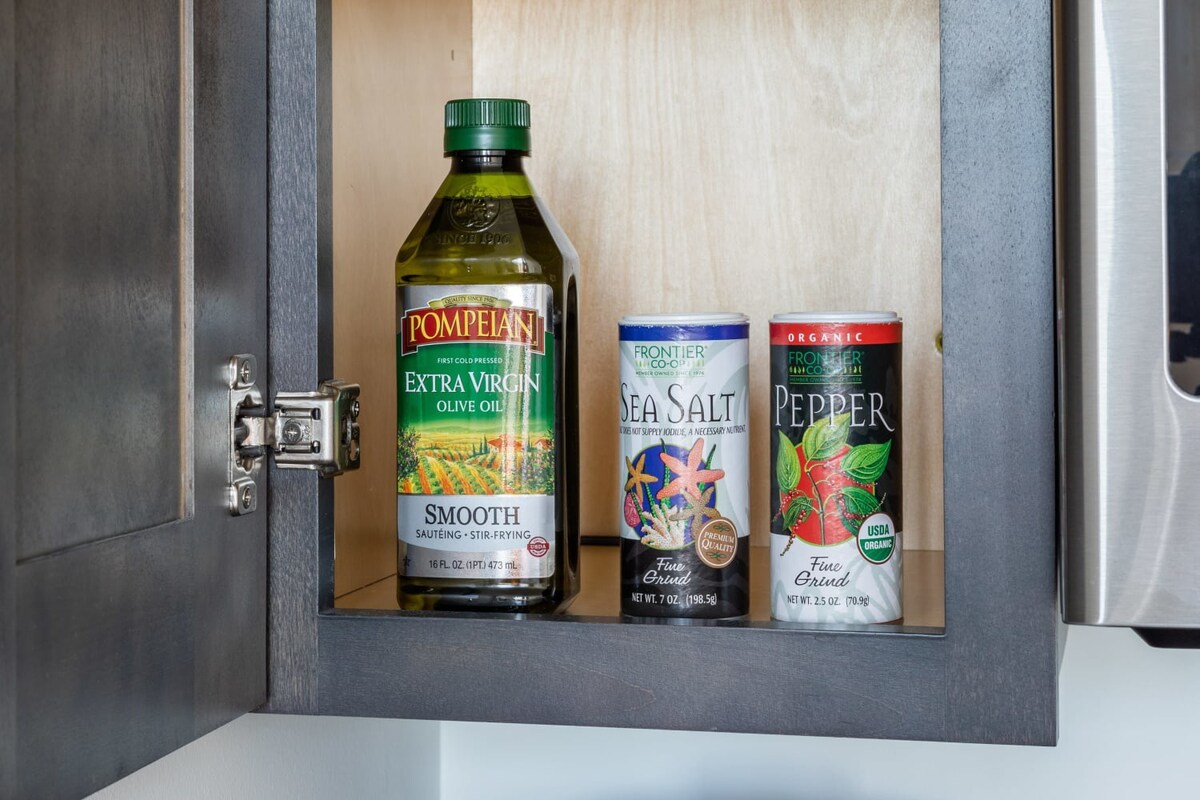 An open kitchen cabinet displaying essential cooking ingredients, including a large bottle of Pompeian Extra Virgin Olive Oil, a container of Frontier Co-op Sea Salt, and a container of Frontier Co-op Pepper.