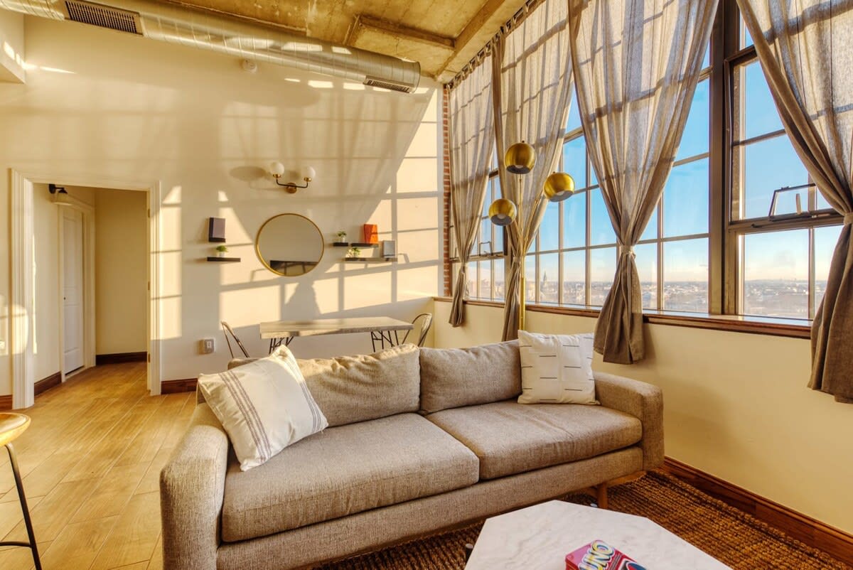 A sunlit living room with a light gray sofa, a white hexagonal coffee table, and expansive industrial-style windows offering city views. The space features high ceilings with exposed concrete and a textured area rug.