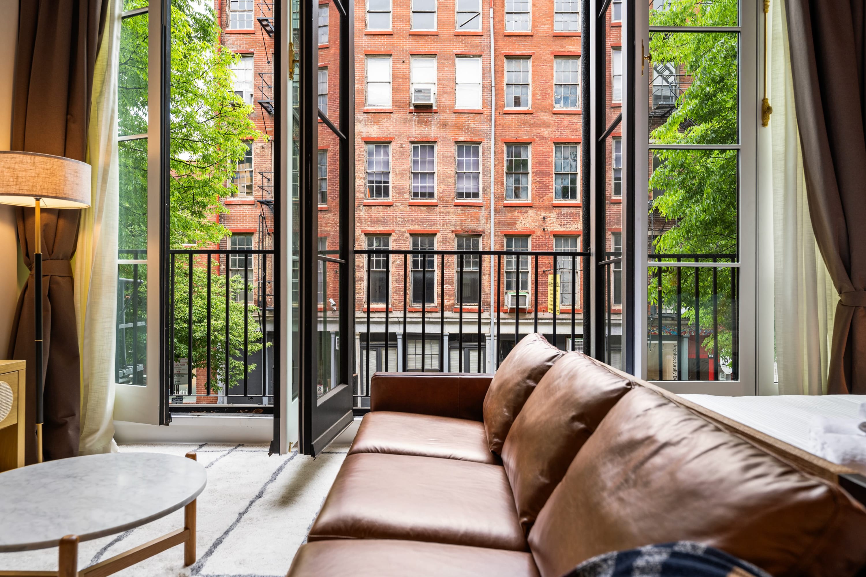 A close-up view from the Juliet balcony, showcasing the classic brick facades of neighboring city buildings with fire escapes and green trees. The black metal railing of the balcony is visible in the foreground.