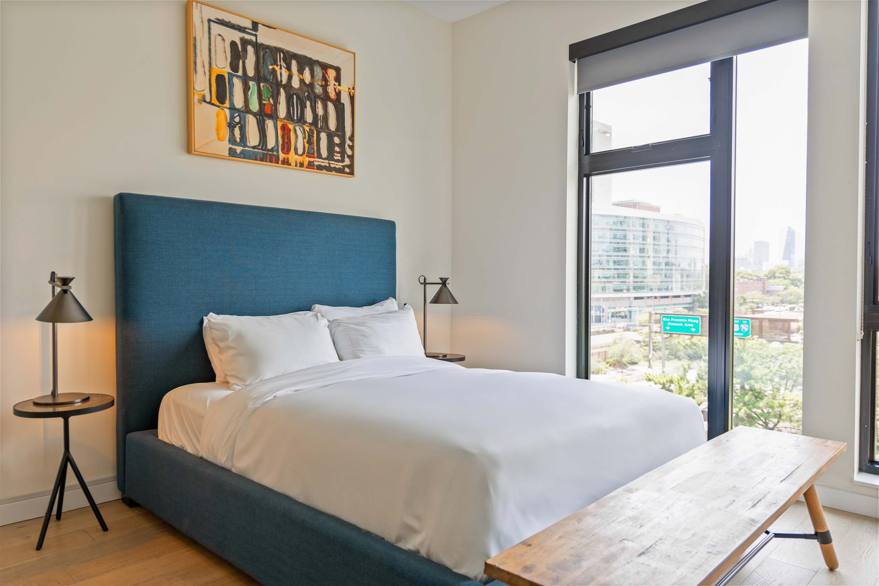 Bright bedroom with a blue upholstered queen-sized bed dressed in white linens, flanked by two black metal nightstands with lamps, and a wooden bench at the foot of the bed. A large window provides a view of the city.
