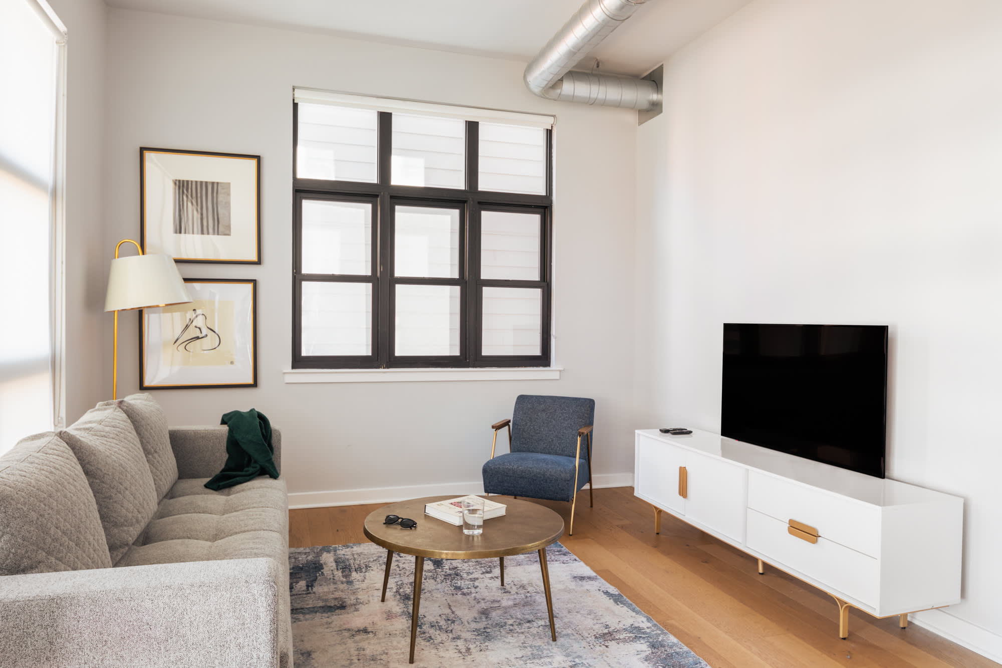 Modern living room with a light gray sofa and a white media console with a TV, facing a round brass coffee table and a blue accent chair. Large windows with blinds provide natural light, and two framed artworks hang on the wall.