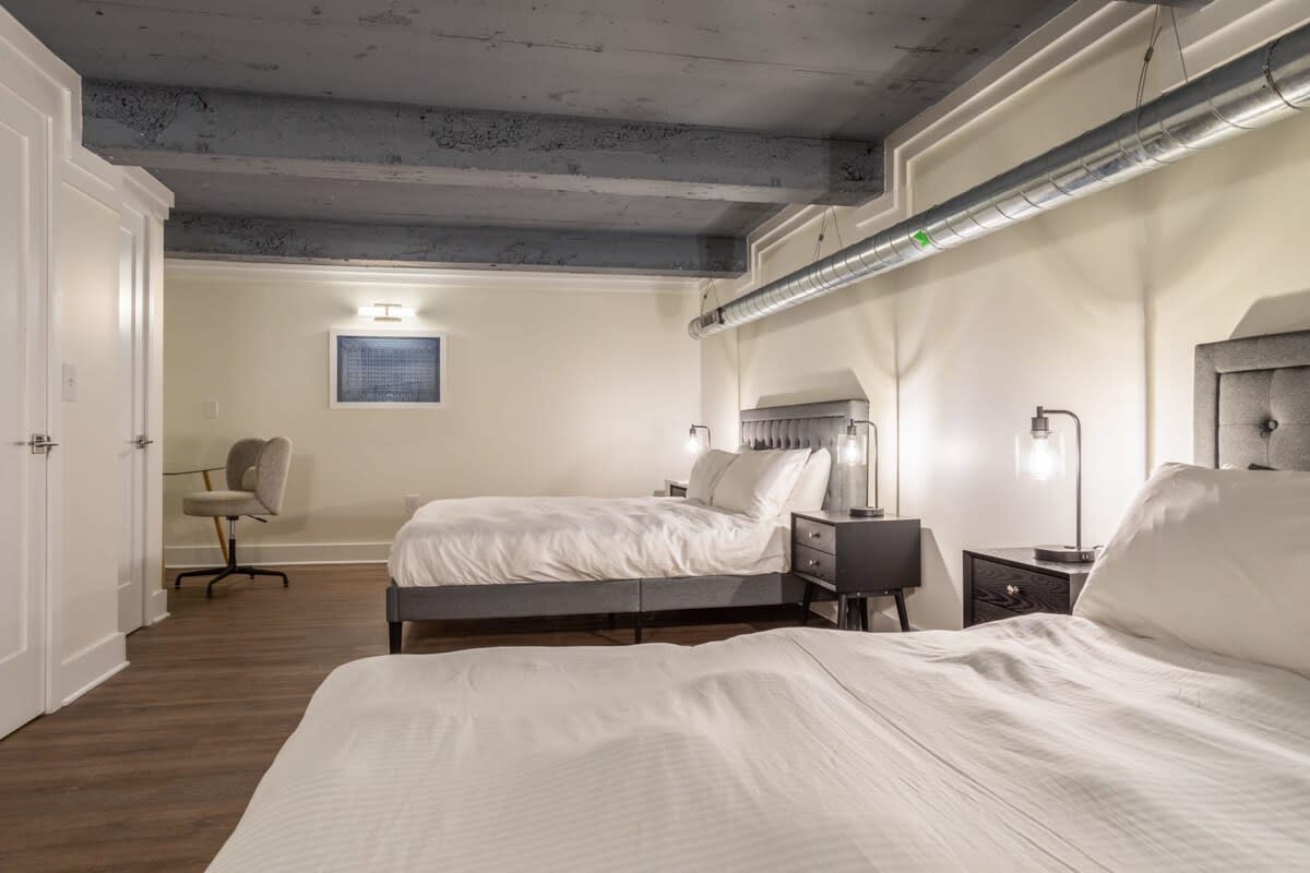 Spacious bedroom-1 featuring two gray upholstered queen beds with white bedding, dark wood nightstands, and industrial-chic exposed ductwork and concrete ceiling beams, with a small office nook.