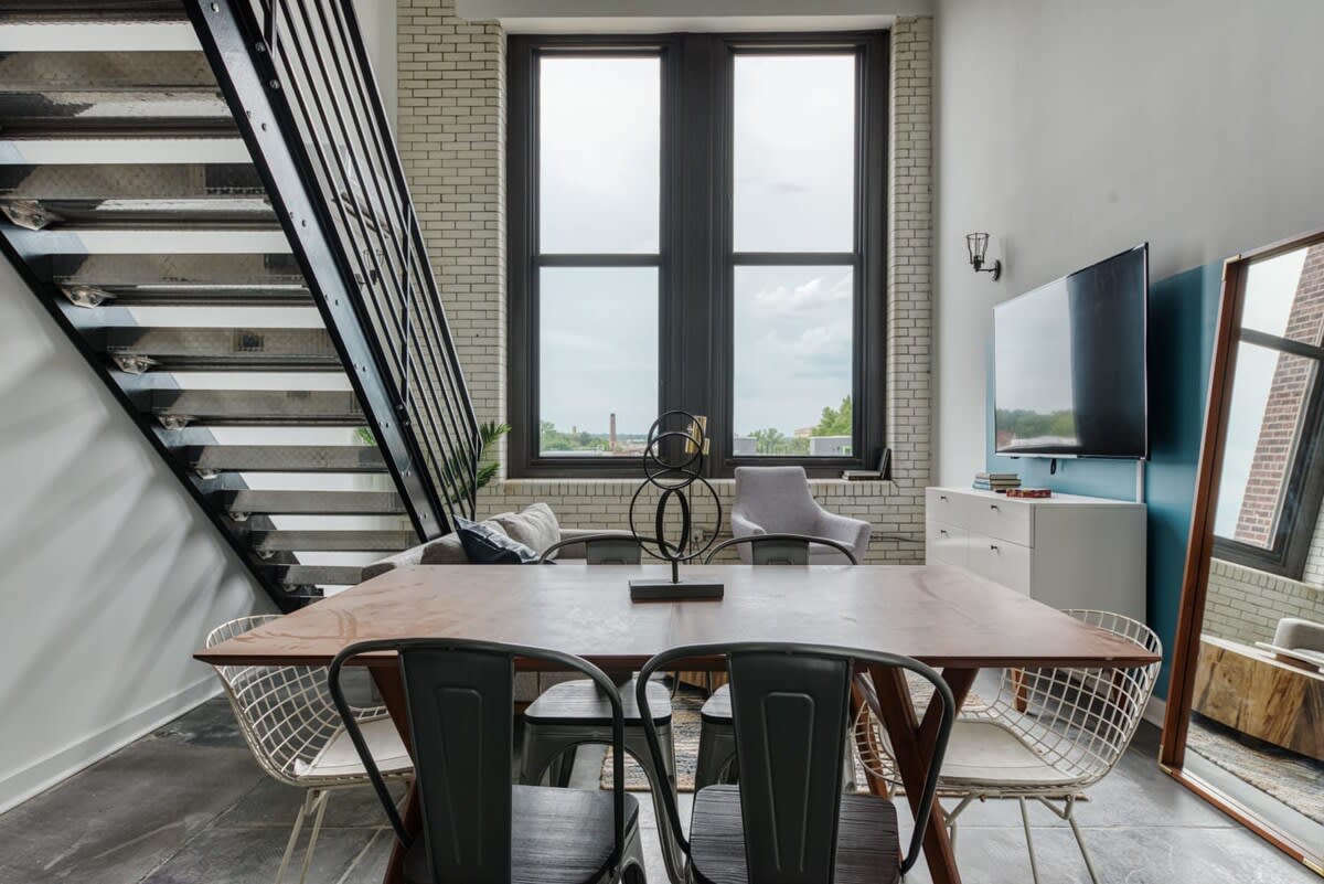 Dining area with a long wooden table and mixed metal and wooden chairs, positioned in front of tall industrial-style windows, adjacent to a modern metal staircase.