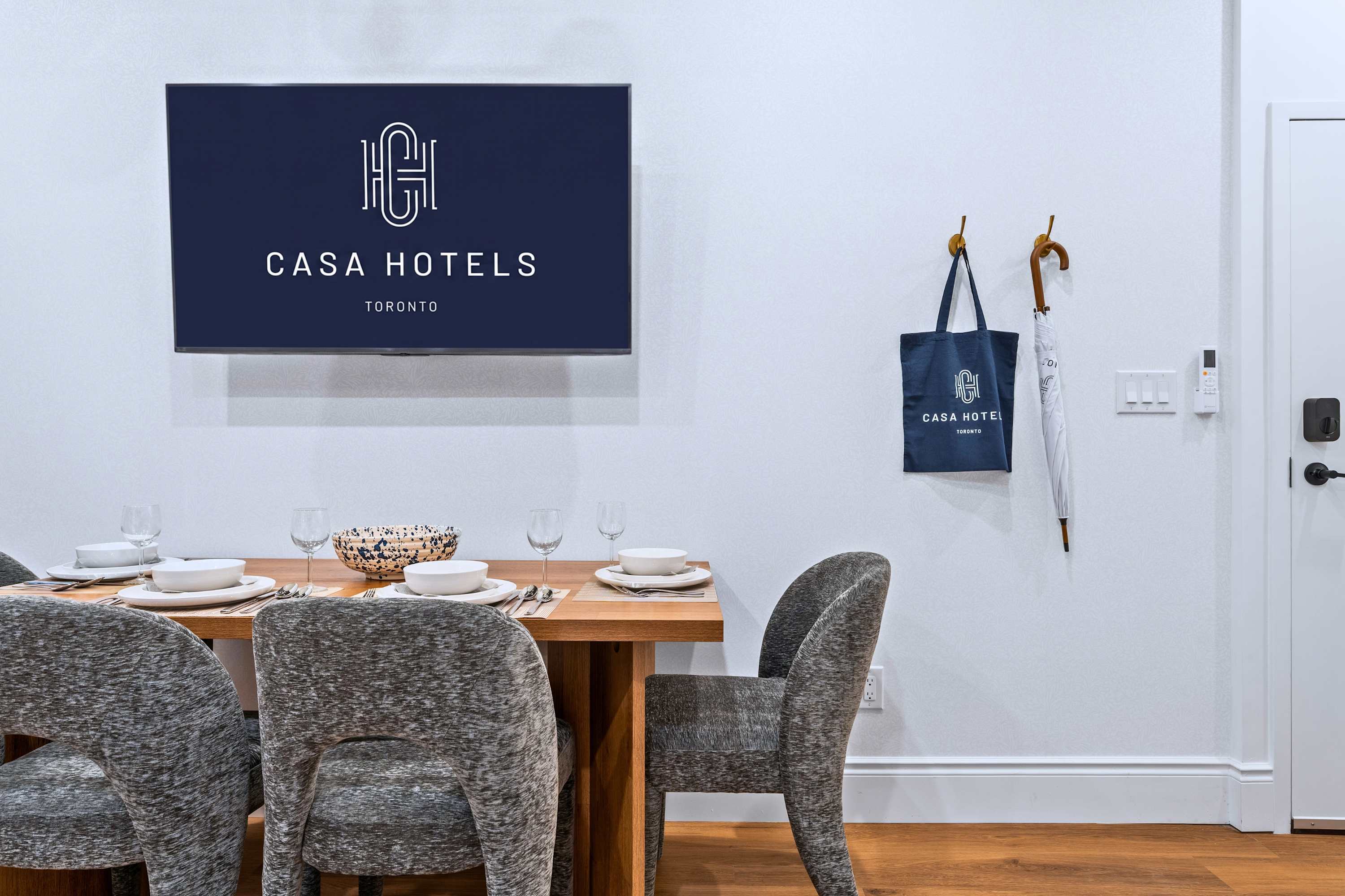 A close-up of the modern dining area shows a light wood table and grey upholstered chairs, with a wall-mounted TV displaying a hotel logo and a blue tote bag hanging on a wall hook next to an umbrella.