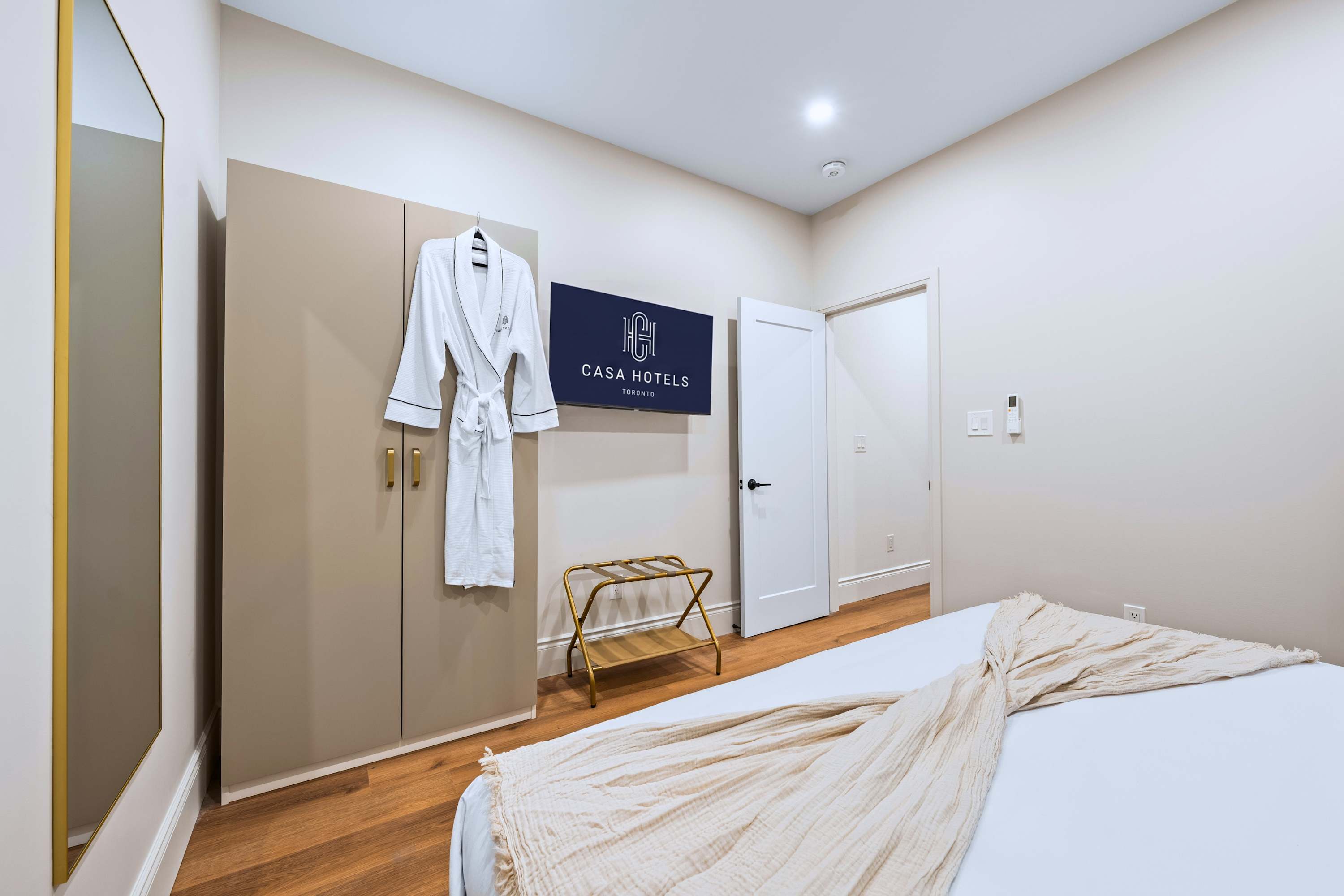 This bedroom includes a king-sized bed, a large wardrobe with gold handles, a wall-mounted TV displaying a hotel logo, and a gold luggage rack, with a white bathrobe hanging on the wardrobe.
