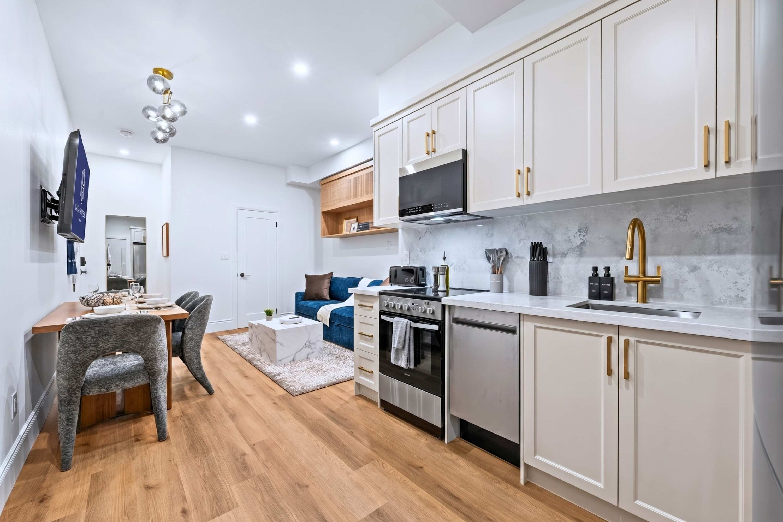 An open-concept living space combines a dining area with a wooden table and grey chairs, a kitchen featuring white cabinets and stainless steel appliances, and a cozy living area with a blue sofa.