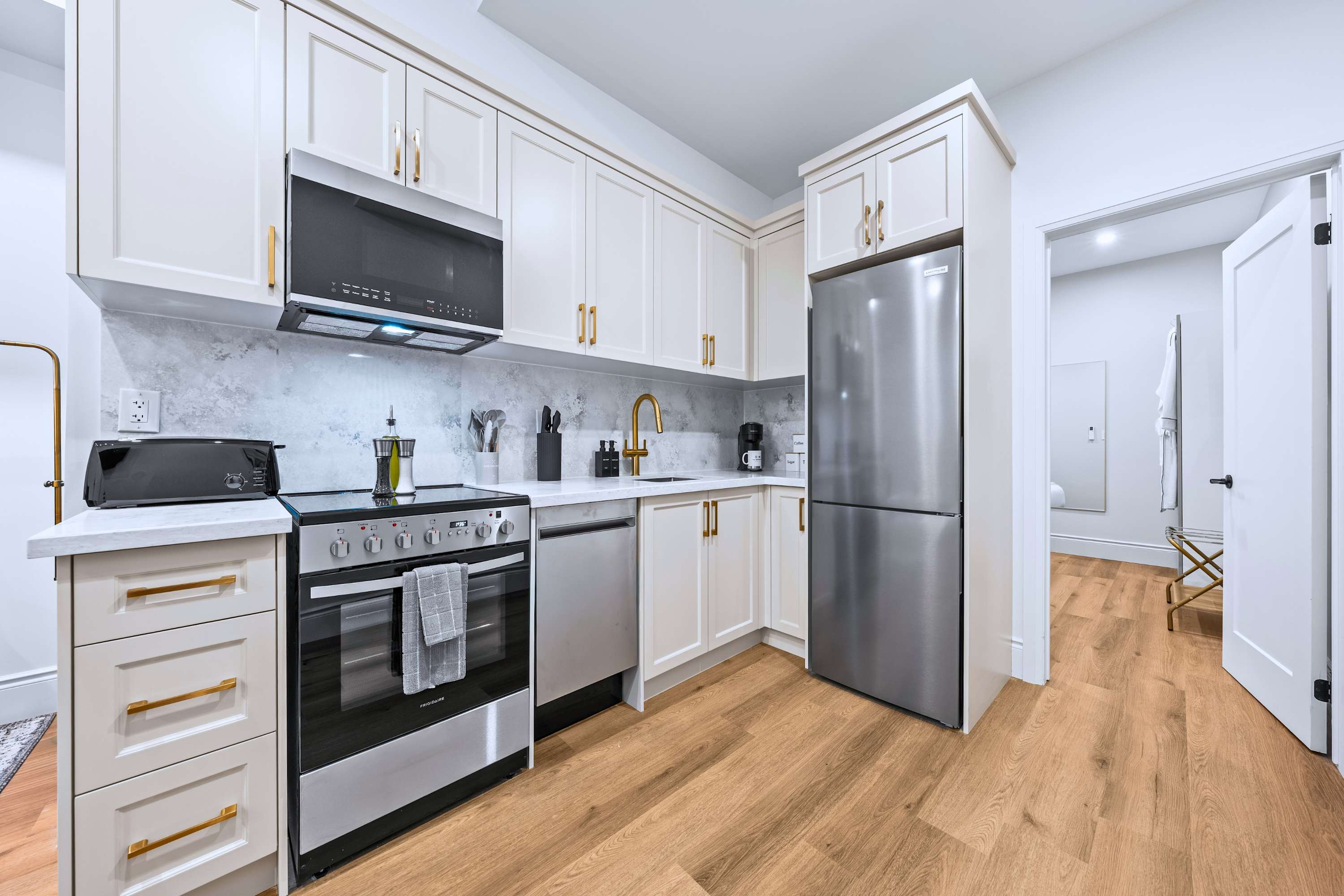 A full view of the modern kitchen reveals white shaker-style cabinets, gold hardware, white quartz countertops, and stainless steel appliances including a refrigerator, range, microwave, and dishwasher, all set on warm hardwood floors.