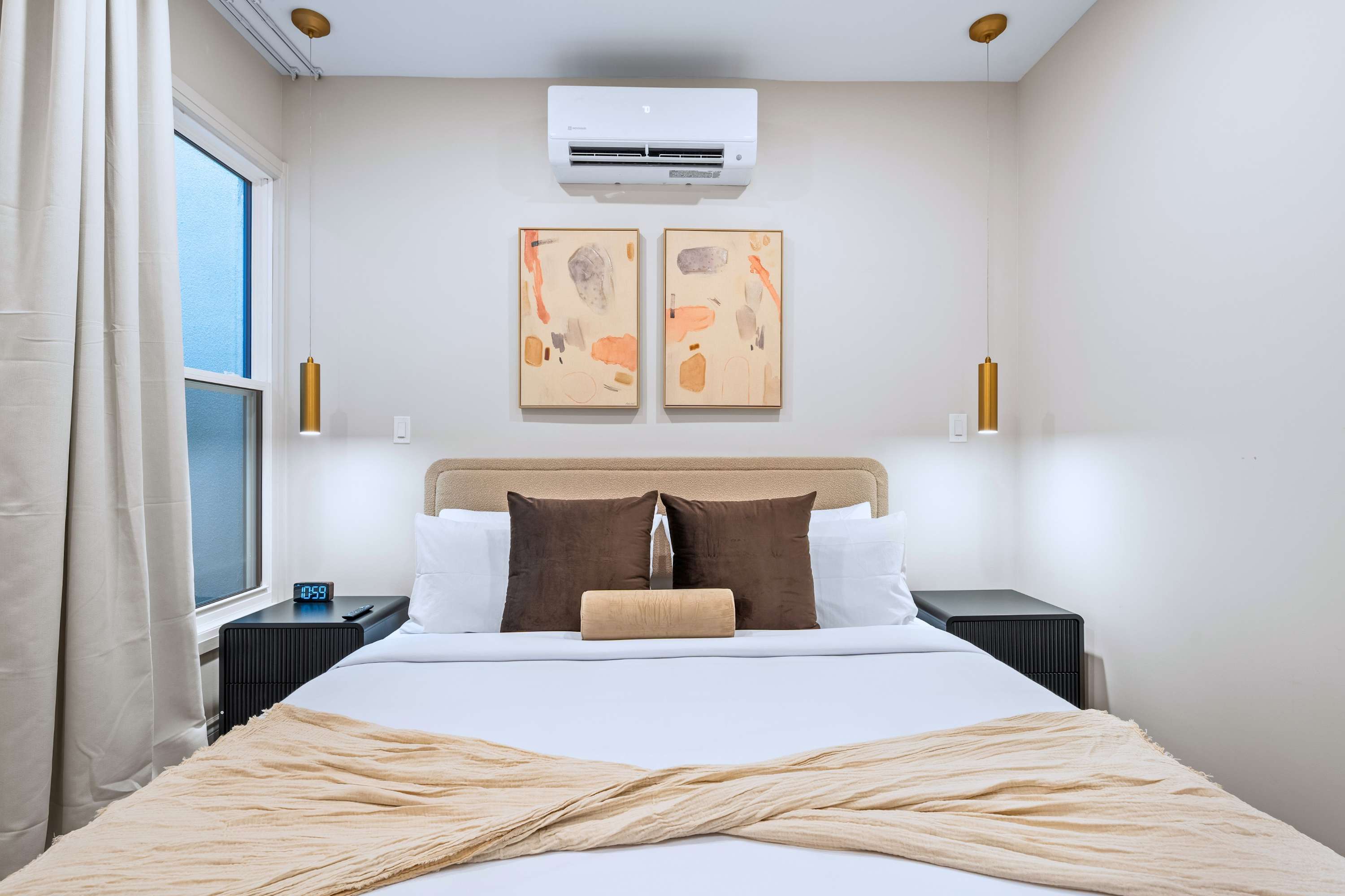 A bright bedroom showcases a king-sized bed with a beige upholstered headboard and black nightstands, complemented by gold pendant lights, abstract artwork, and a window with light-colored curtains.