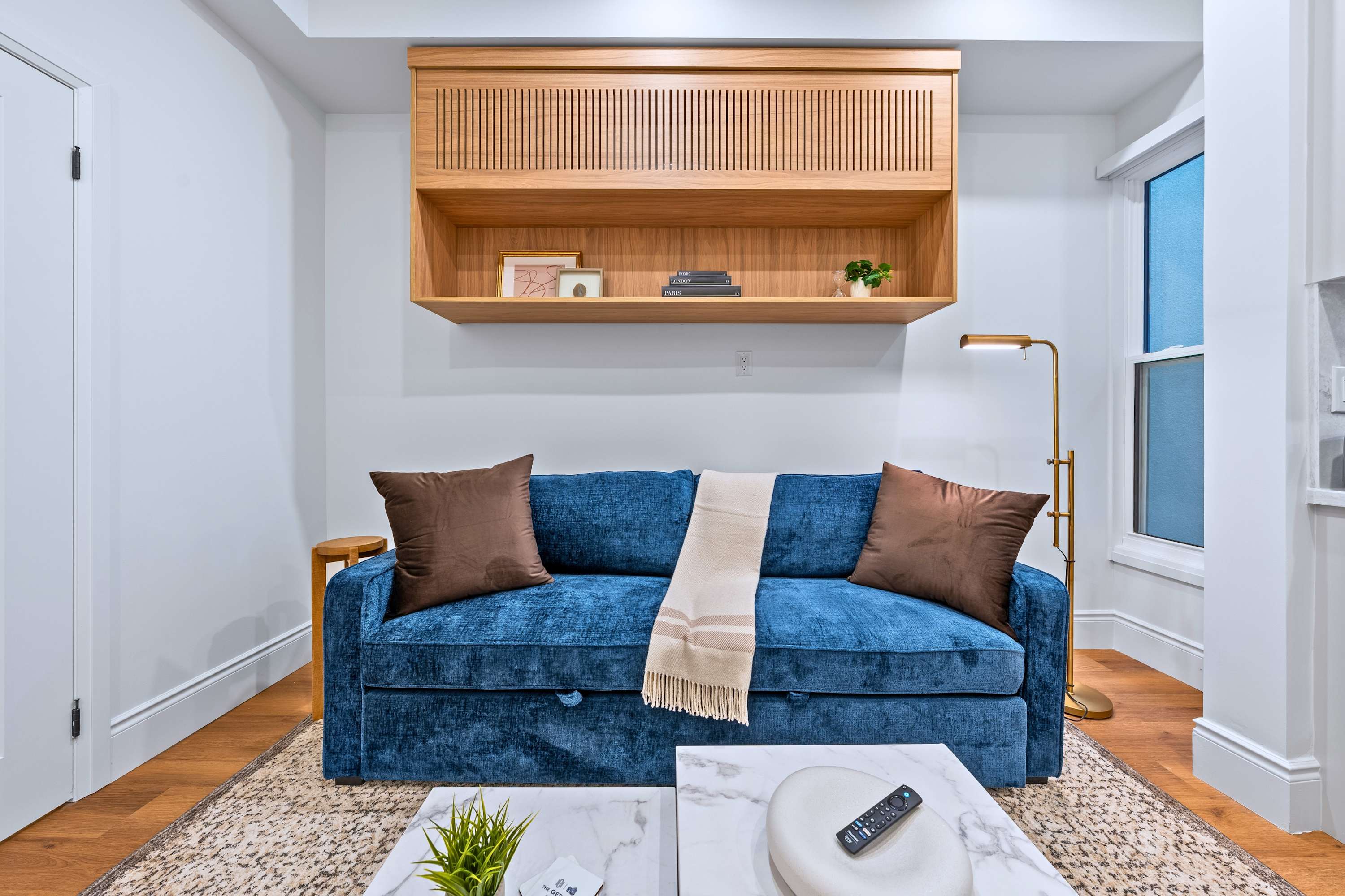 Stylish living room featuring a plush blue velvet sofa adorned with brown pillows and a throw blanket, two white marble coffee tables, and a wooden slatted wall-mounted shelf with decorative items, illuminated by a floor lamp.