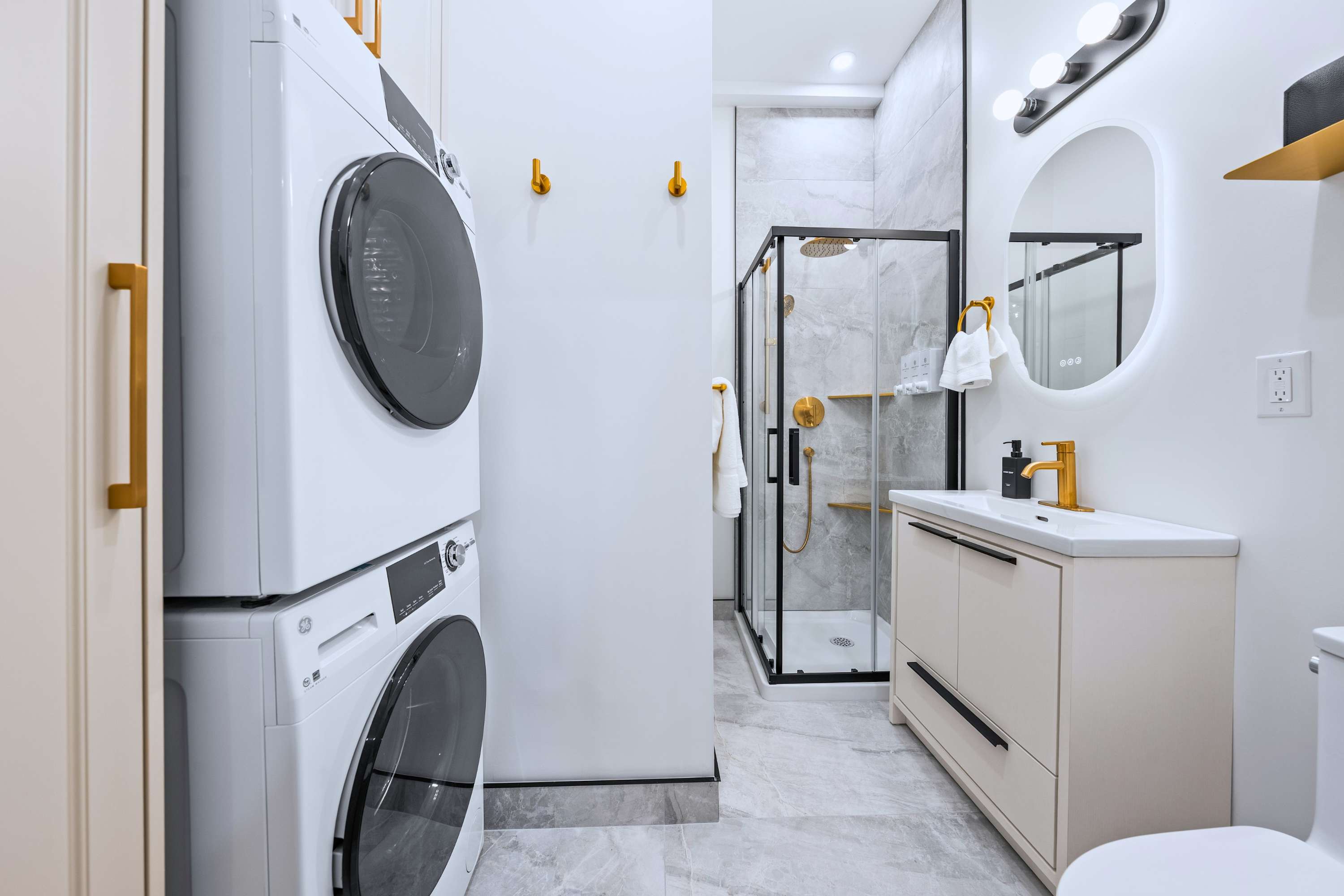 A modern bathroom features a stacked washer and dryer unit, a vanity with a white sink and gold faucet, a round mirror, and a glass-enclosed shower with black framing and gold fixtures.