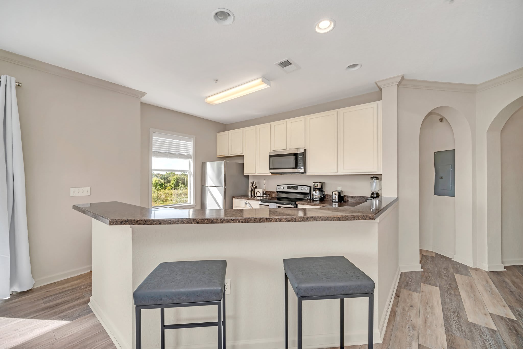 Well-equipped kitchen featuring light cream shaker-style cabinets, dark granite countertops, and stainless steel appliances including a refrigerator, microwave, and electric range. A breakfast bar with stools is visible on the right.