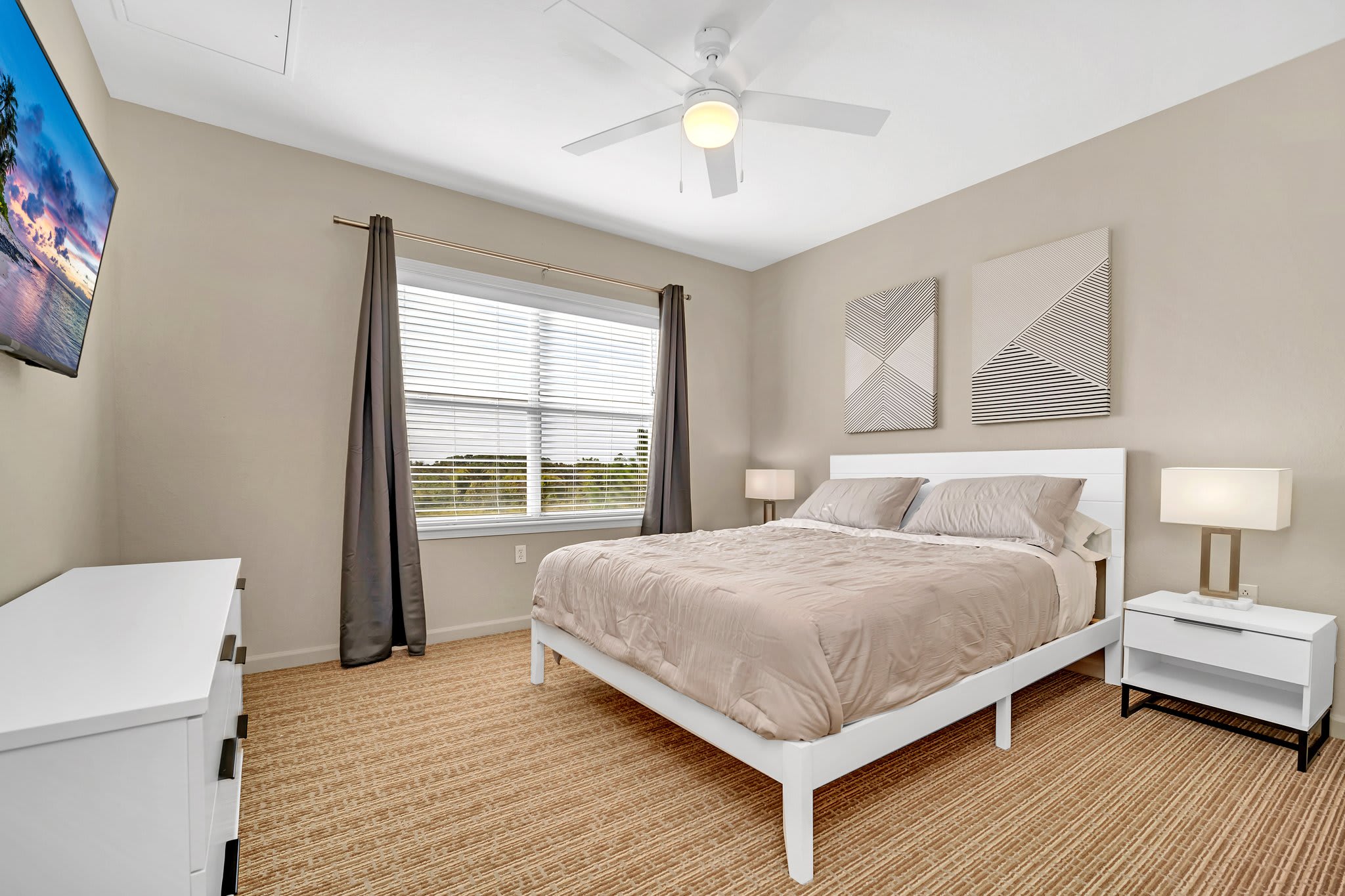 A cozy bedroom featuring a white queen-sized bed with beige bedding, two white nightstands with lamps, a wall-mounted TV, and a ceiling fan, all on a textured carpeted floor.
