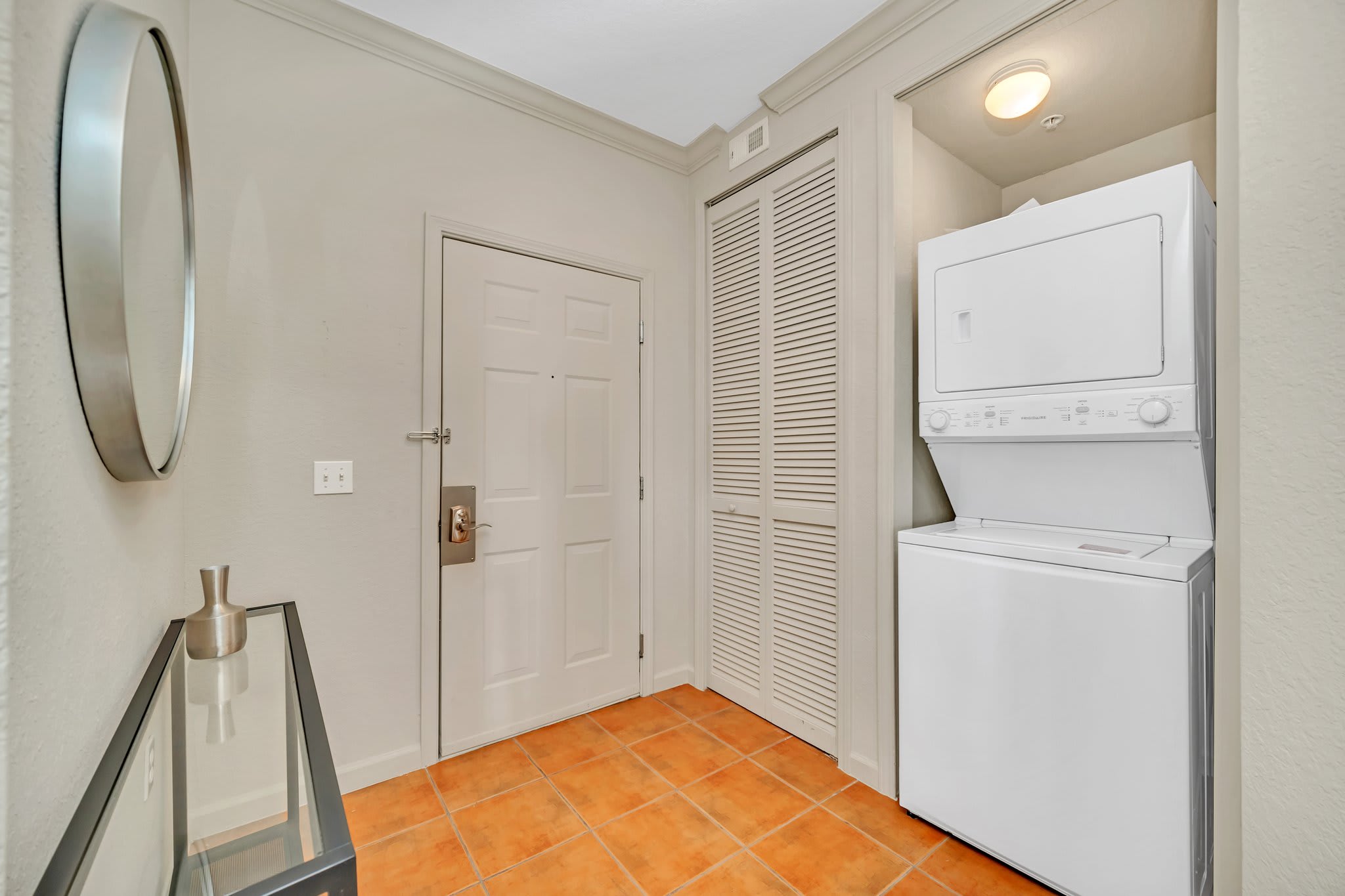 A functional laundry area located in the entryway, featuring a white stacked washer and dryer unit next to a white door, with terracotta-colored tiled flooring.