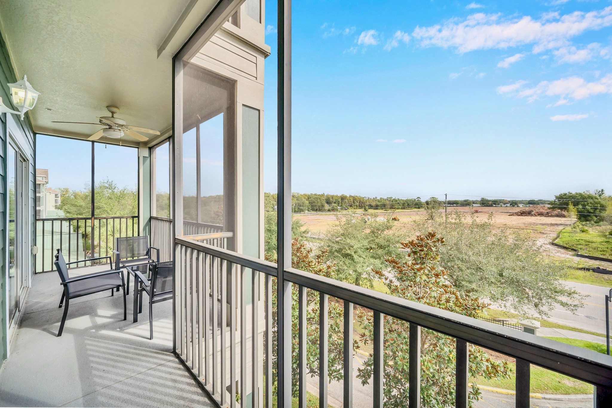 Expansive screened balcony with outdoor seating, offering a panoramic view of a mix of trees and undeveloped land under a bright blue sky. The metal railing and screening provide a secure outdoor space.