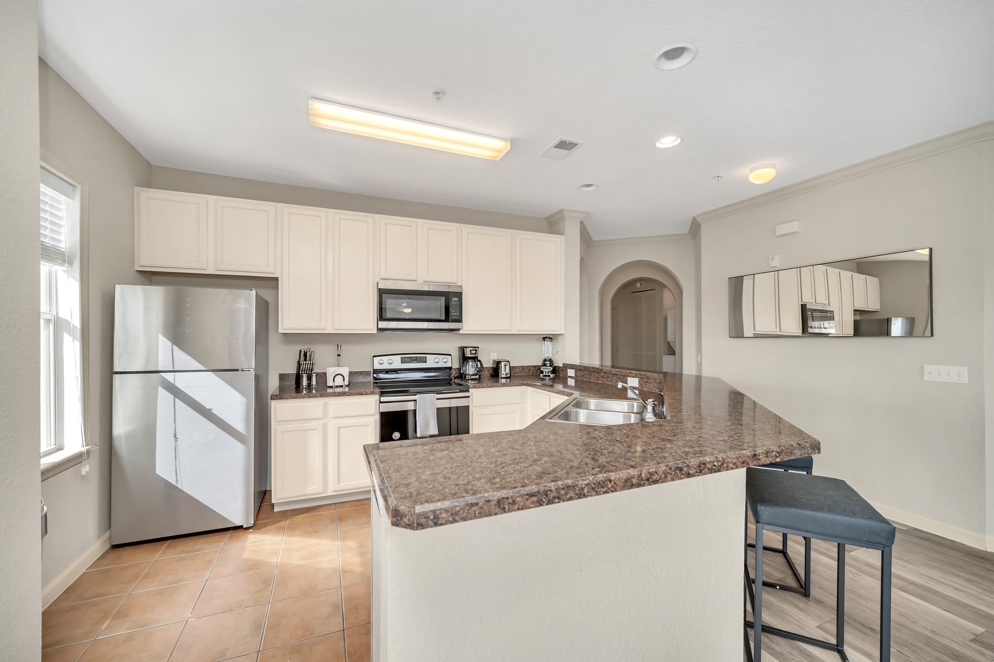 Bright kitchen with light cream cabinetry, dark speckled countertops, and stainless steel appliances. A large breakfast bar with two stools provides casual dining space, and a window offers natural light.
