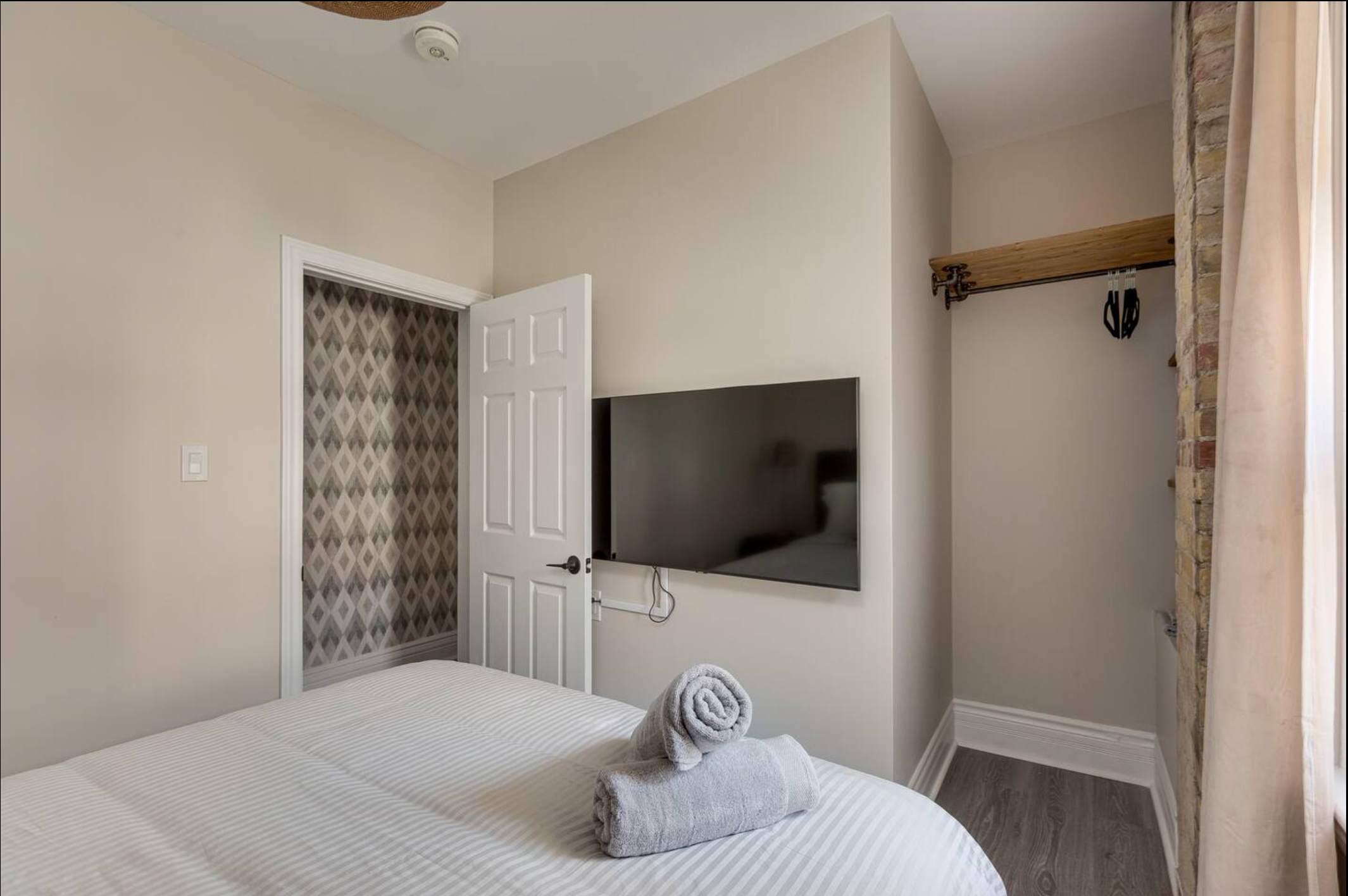 View of bedroom-2 showing a wall-mounted flat-screen TV opposite the bed, a closet with a white bi-fold door, and an exposed brick accent wall on the right. Rolled towels are on the bed.