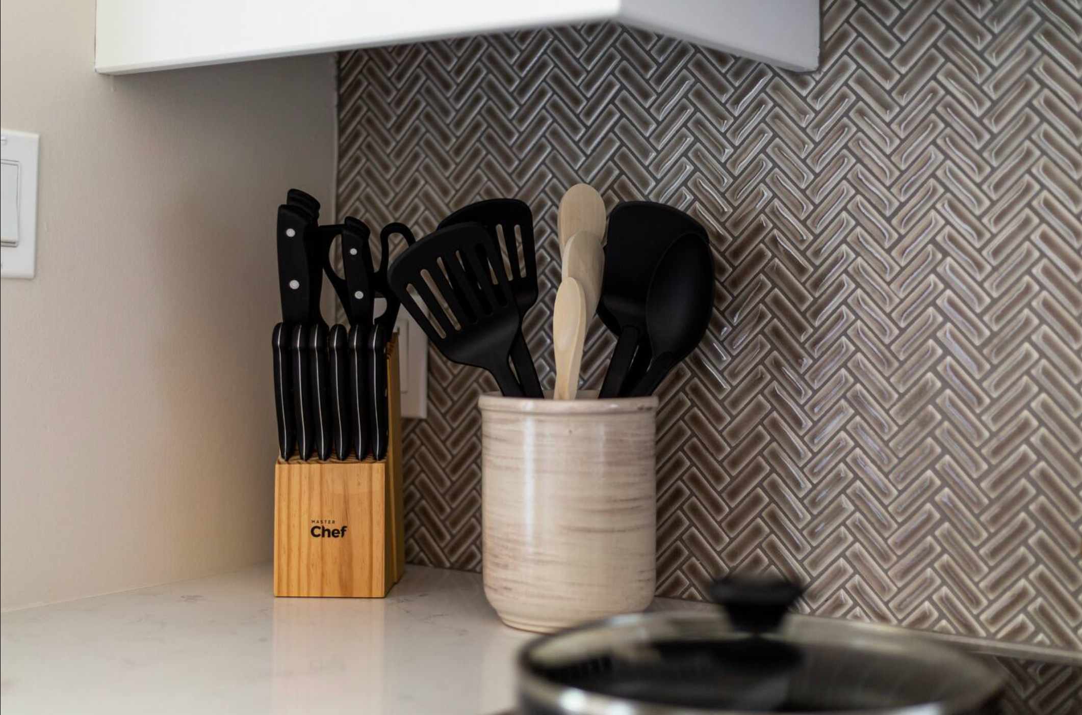 Close-up of a kitchen countertop featuring a wooden knife block with black-handled knives and a ceramic crock holding black and wooden cooking utensils, set against a herringbone tile backsplash.